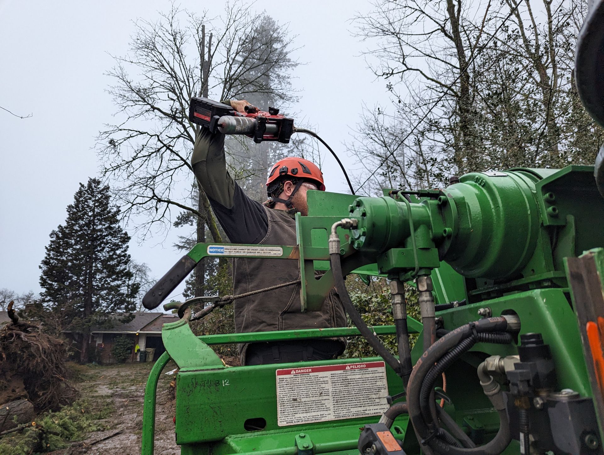 Worker in orange safety gear using a chainsaw on a tree beside green forestry machinery outdoors