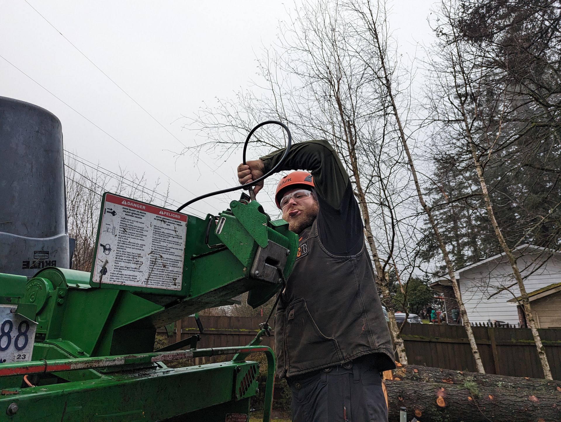 Worker in orange hard hat using a chainsaw beside green machinery outdoors on an overcast day