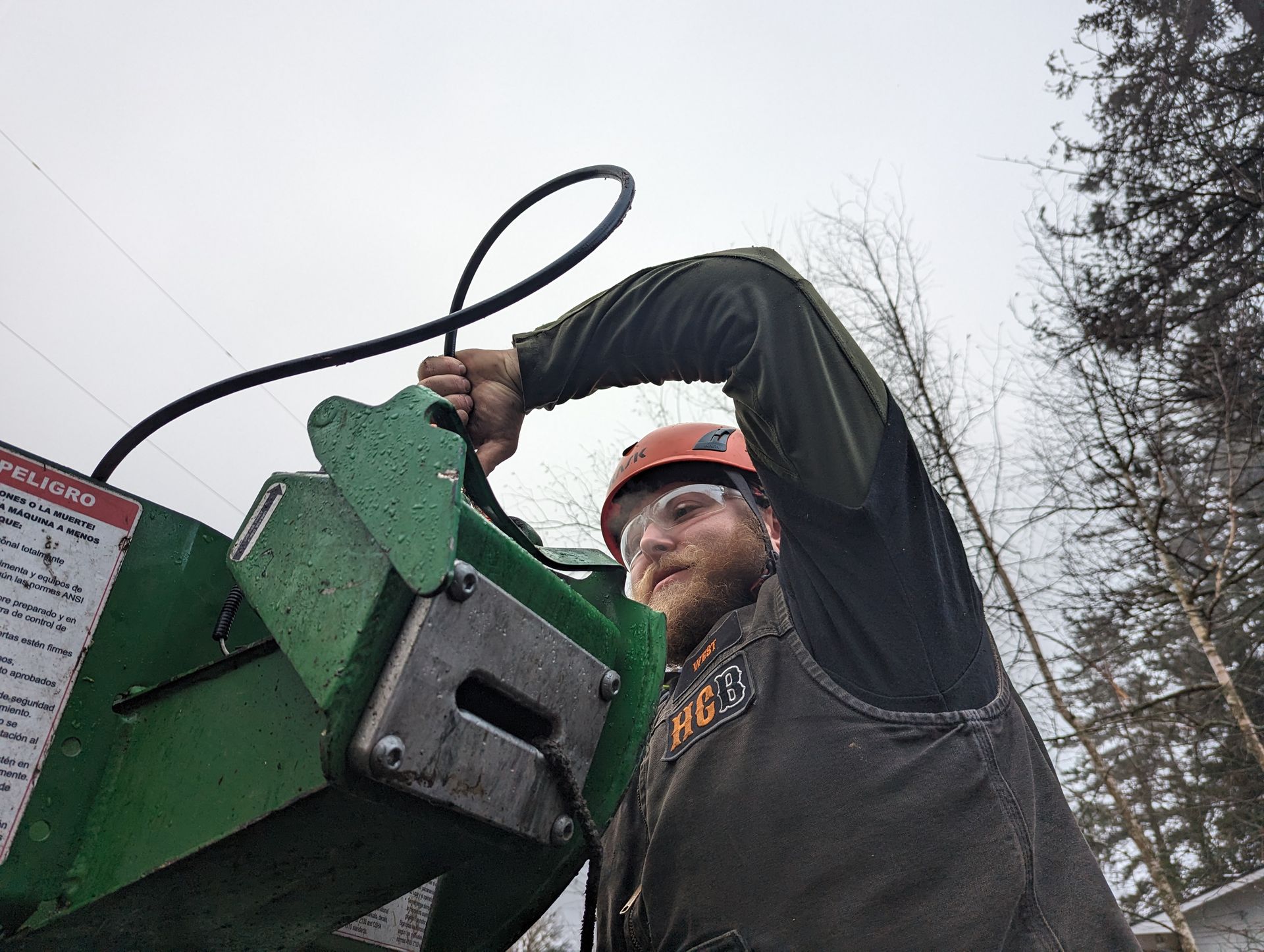Worker in hard hat using a green wood chipper outdoors in snowy weather