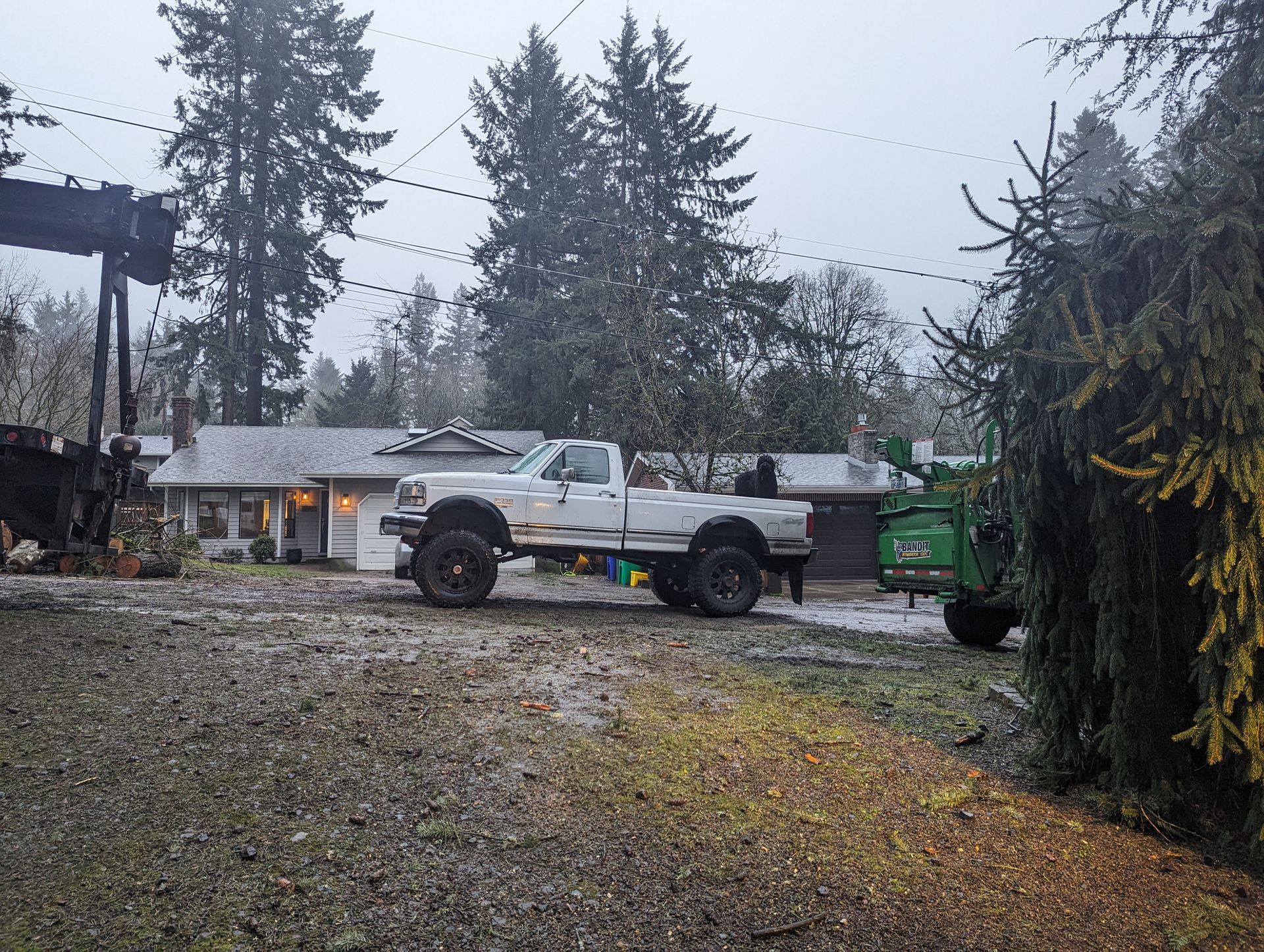 White flatbed truck parked on a wet gravel driveway beside a green dumpster and house, under gray skies