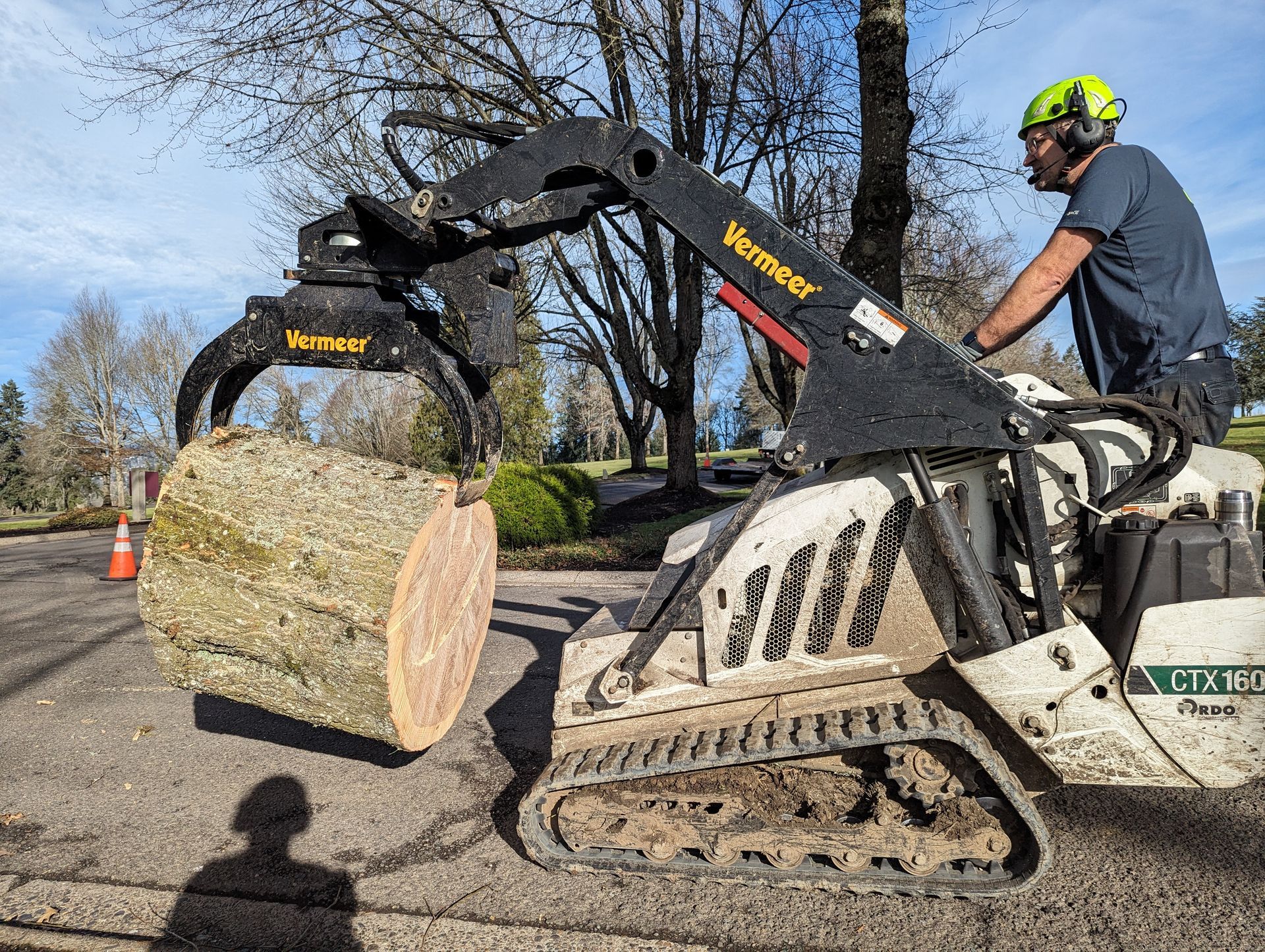 Worker on a tracked loader lifting a large log beside a road under a clear sky.