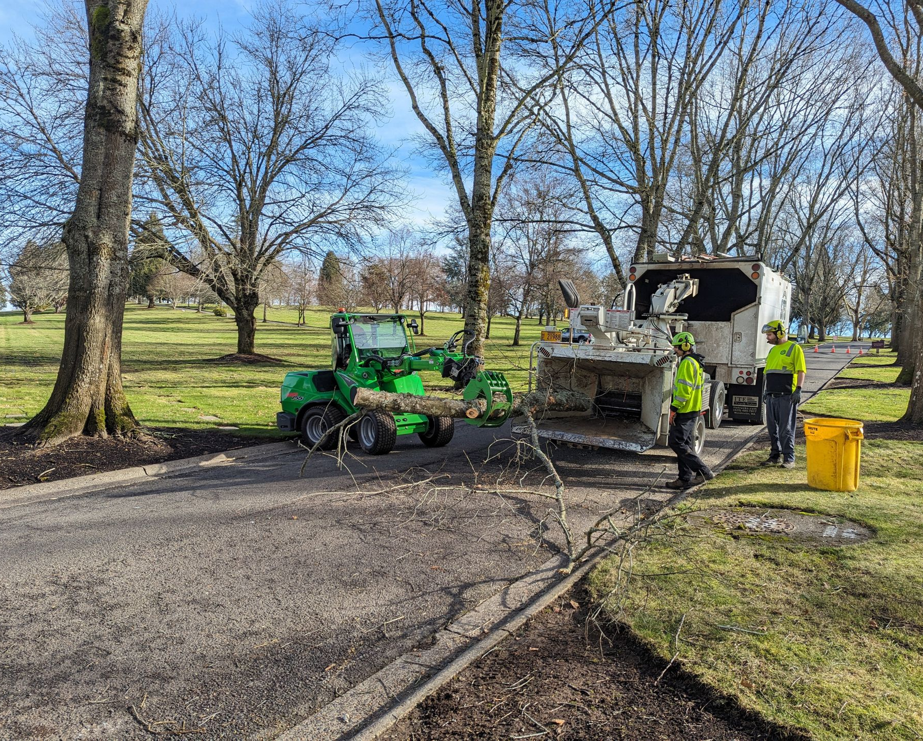 Workers operating green utility equipment on a park path near a truck and trees