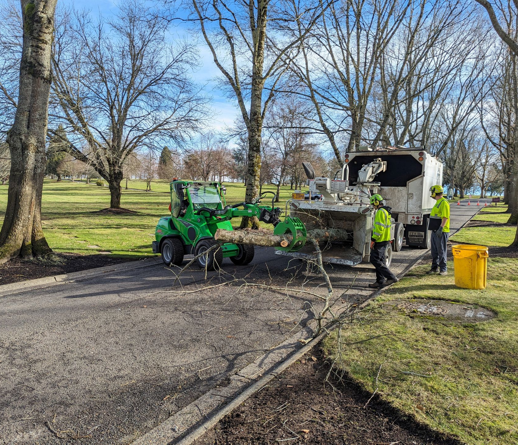Green tractor and white utility truck on a paved path in a park, with trees and grass in the background.