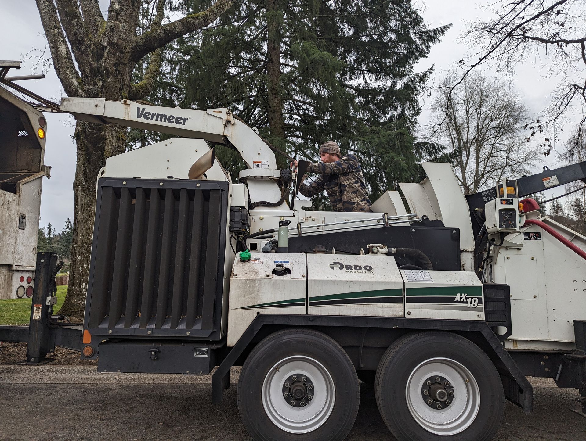 White Woodchipper truck with attached loader arm parked on a street beside a house and trees