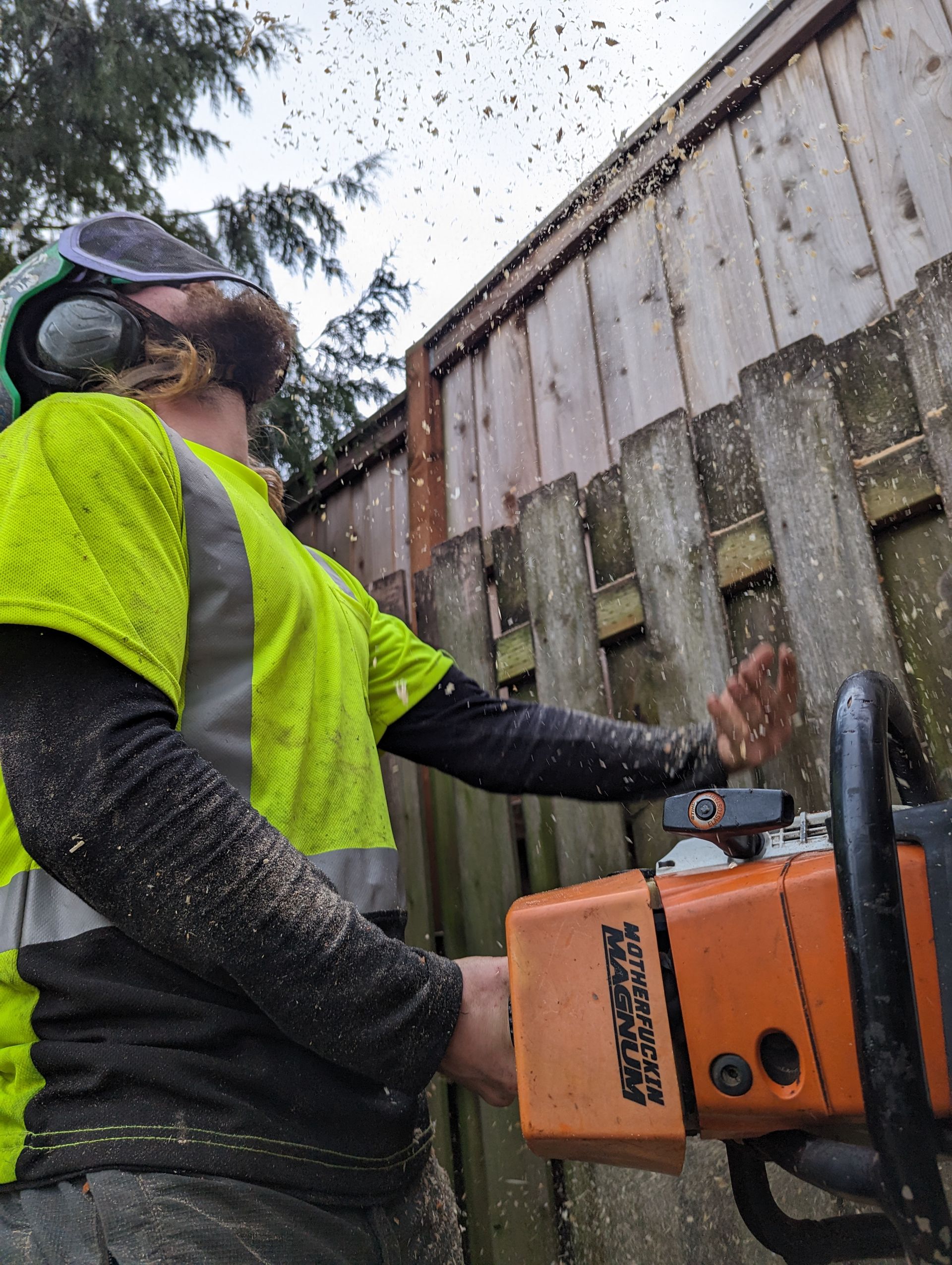 Worker in neon yellow jacket using a chainsaw beside a wooden fence outdoors