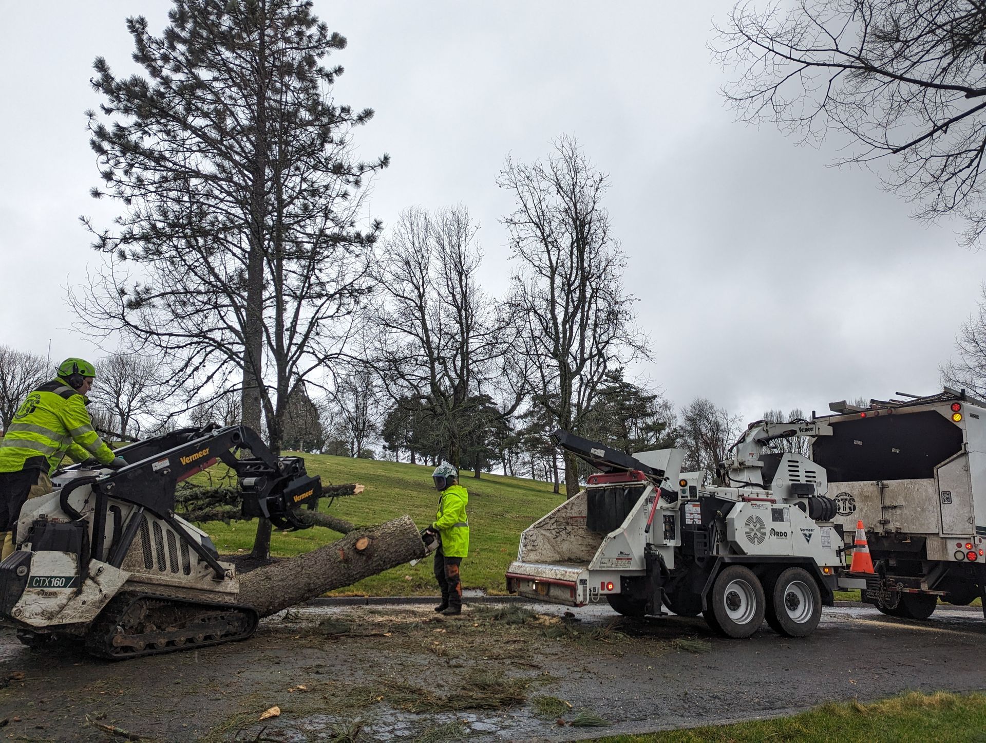 Road crew loading rubble into a dump truck with an excavator on a wet roadside under gray skies