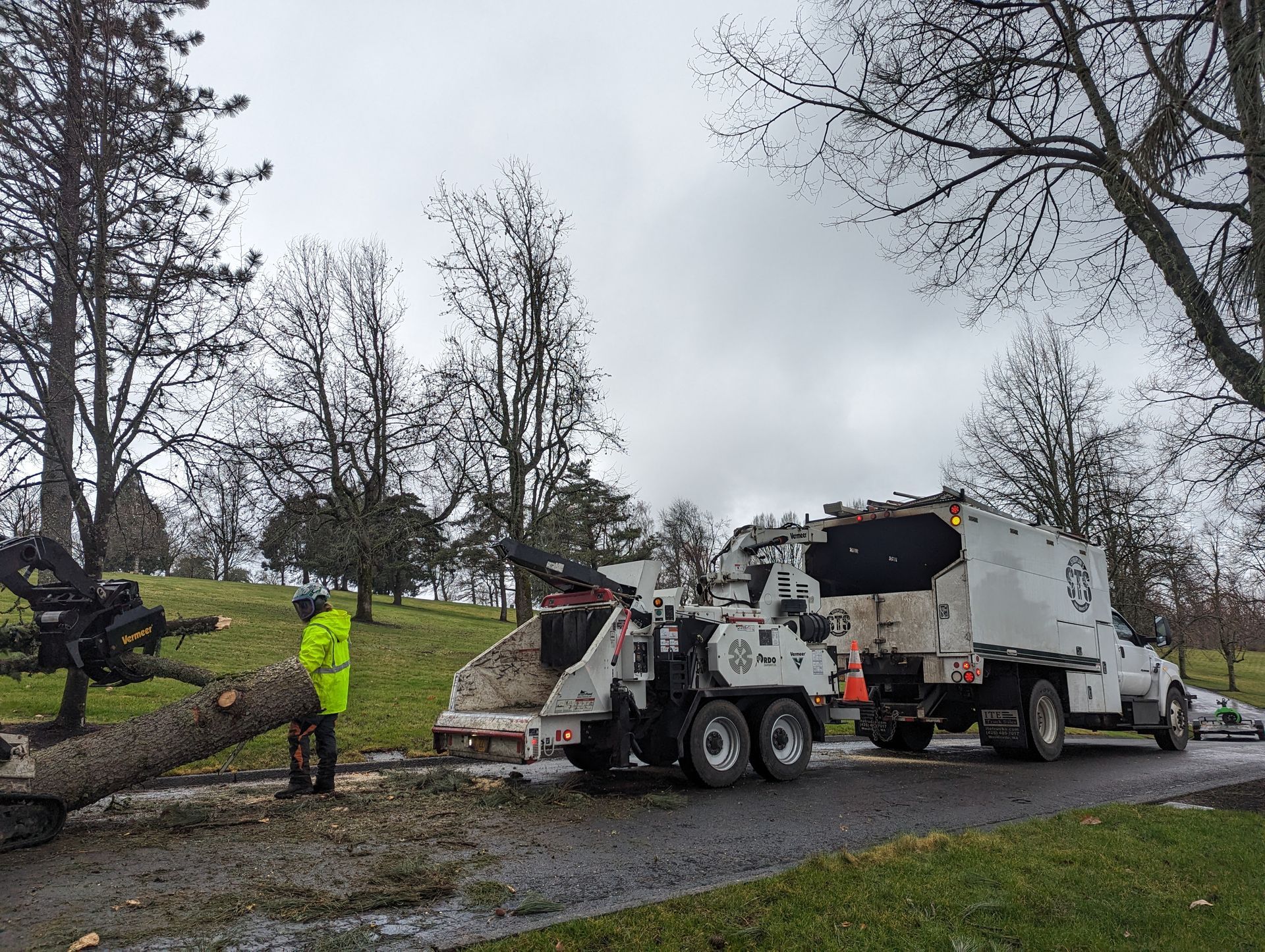 Workers and a utility truck clearing debris on a wet roadside under cloudy skies