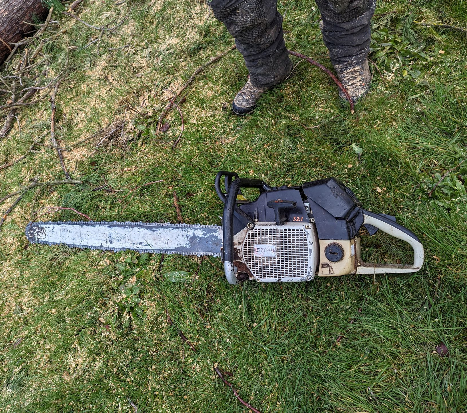 Chainsaw lying on grass beside a person’s boots, outdoors.