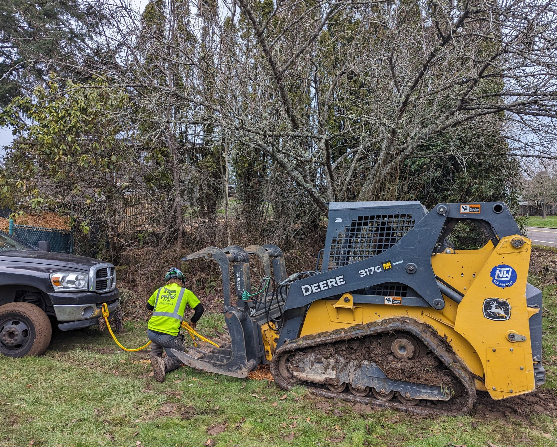 Worker in neon jacket clears debris from a gray-and-yellow skid steer beside a truck in a wooded area