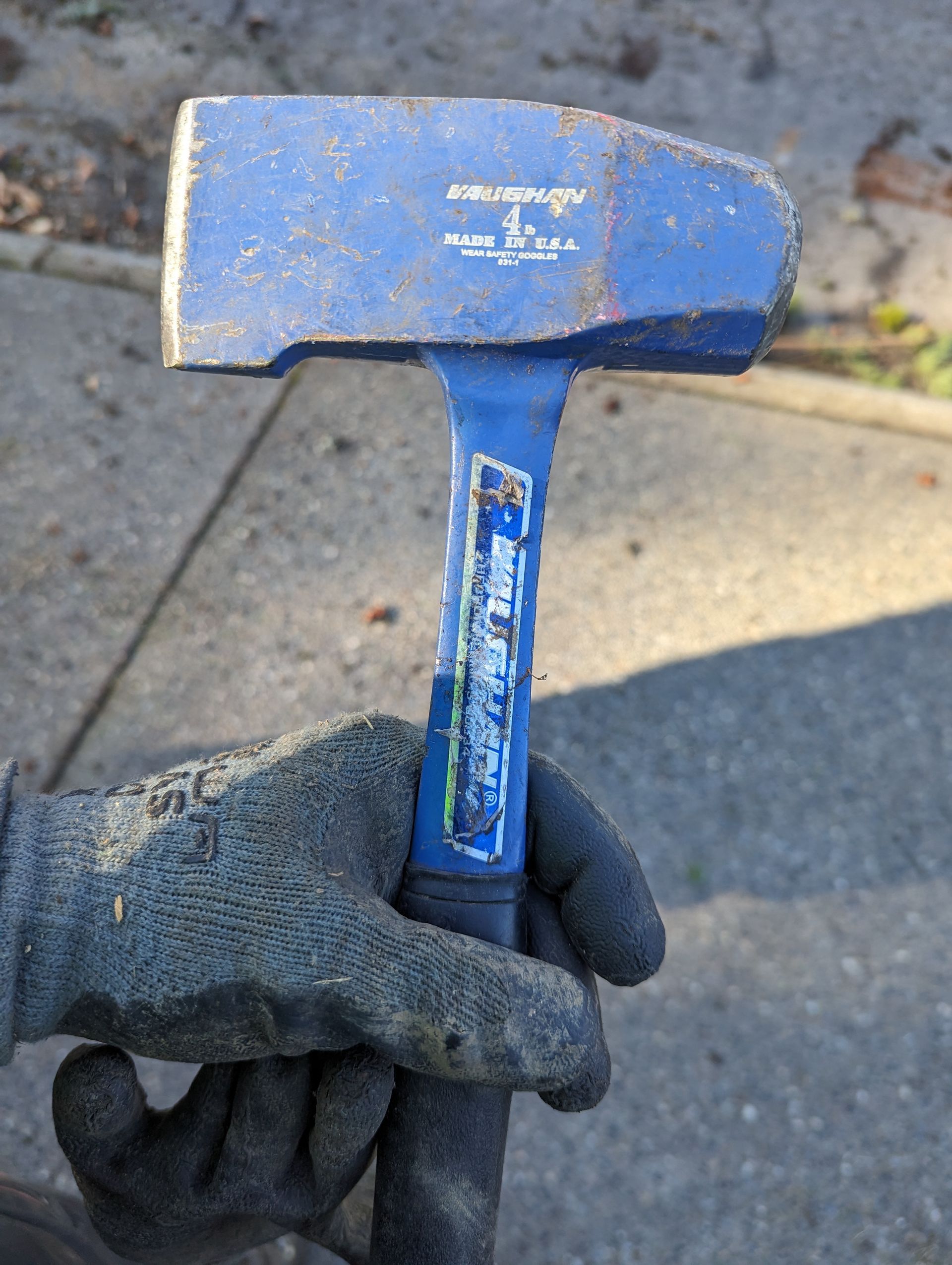 Gloved hand holding a blue hammer with a worn metal head outdoors on pavement