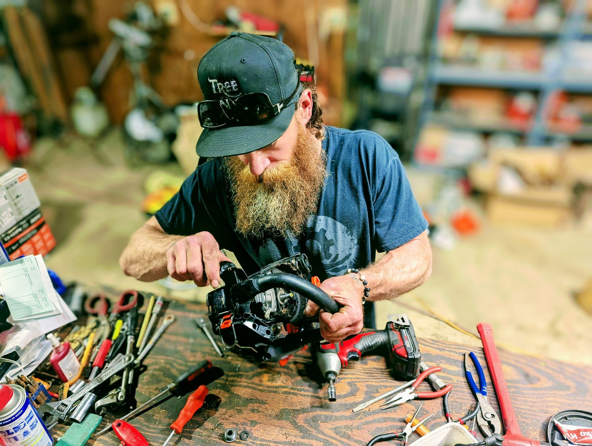 Man repairing a black device at a cluttered workbench in a workshop, with tools spread around him.