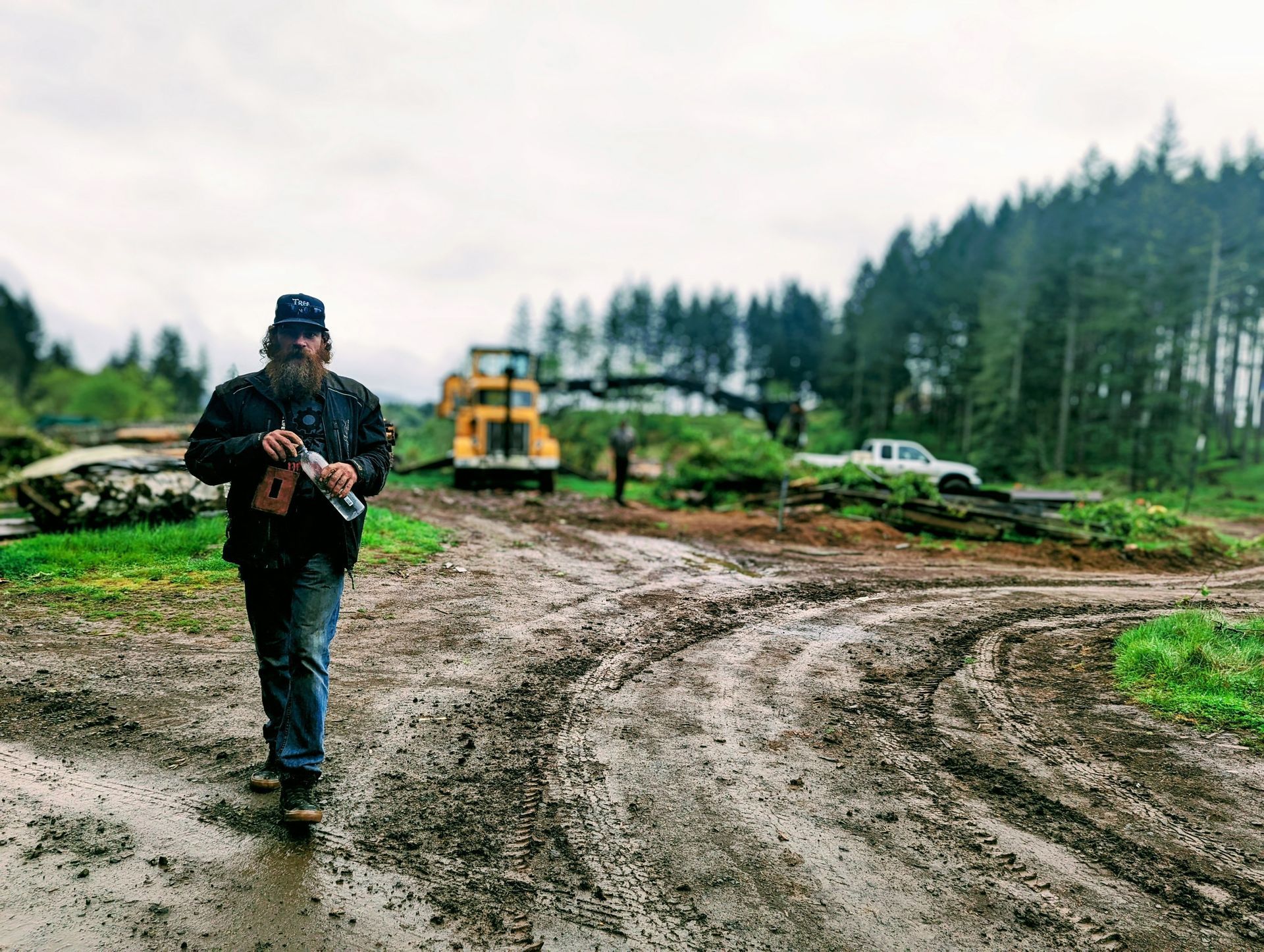 Person walking on a muddy dirt road at a construction site with heavy machinery and trees in the background