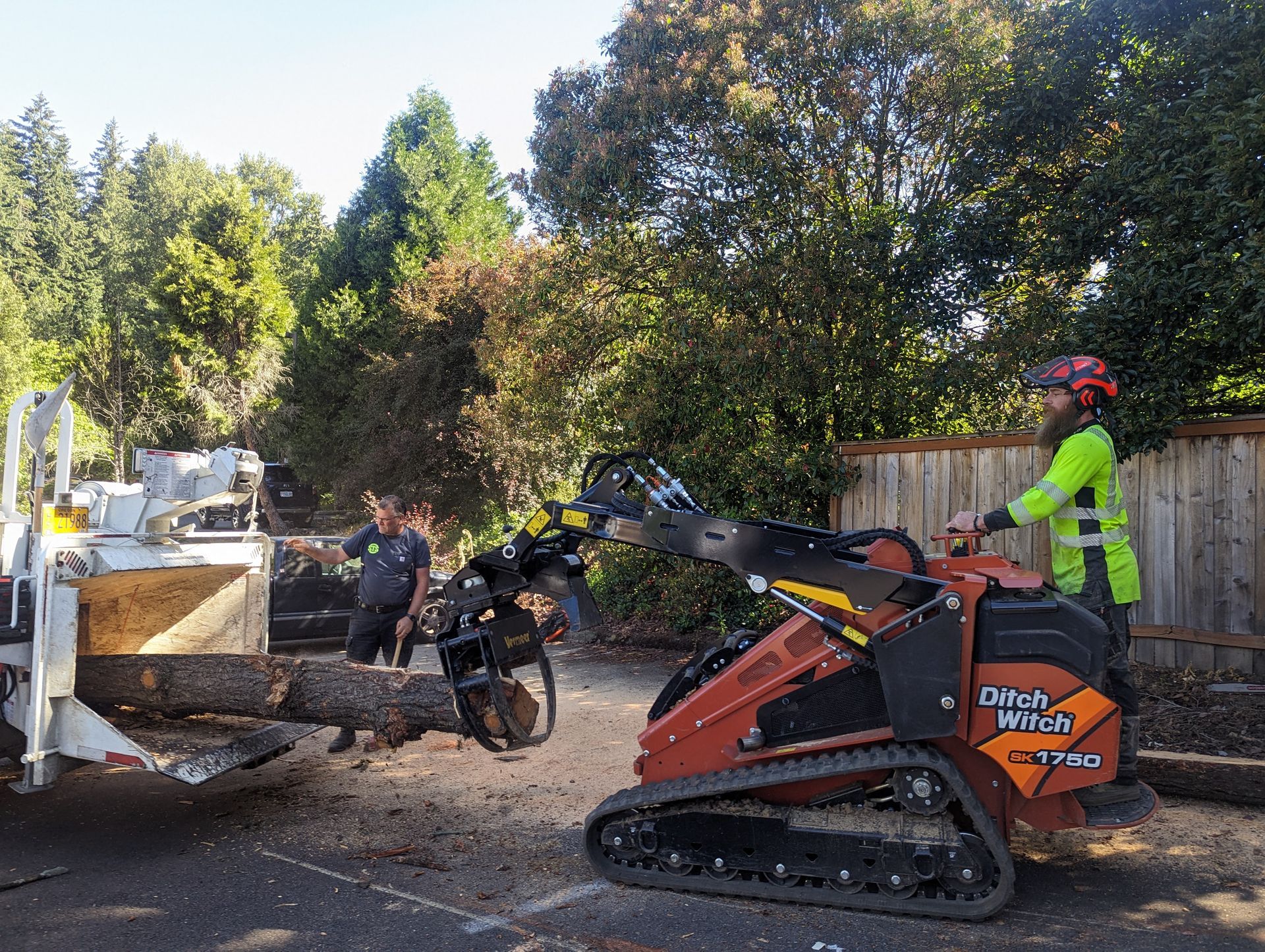 Worker operating an orange mini skid steer at a roadside cleanup site, with another person nearby