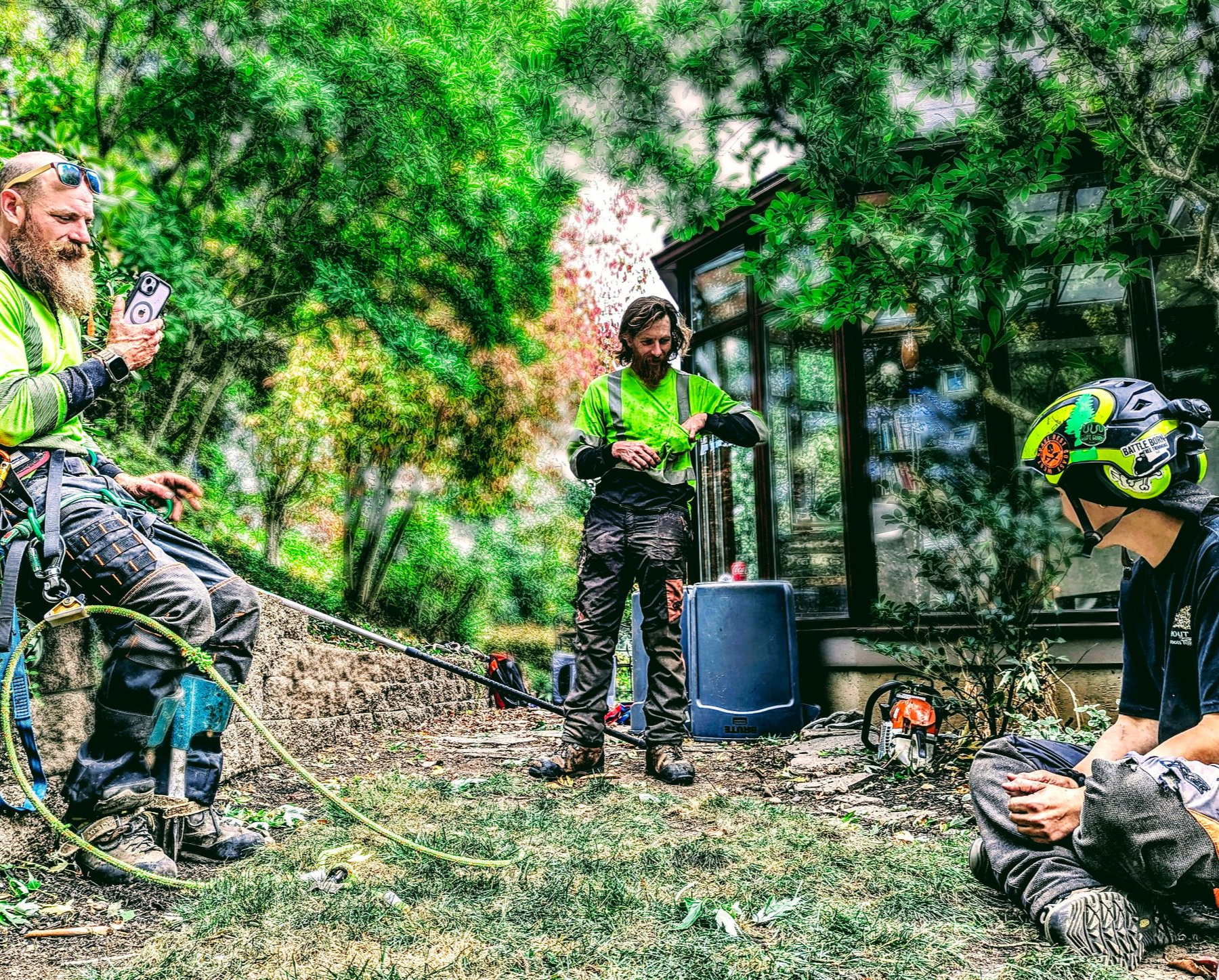 Three people in bright gear outdoors, gathered around equipment in a wooded area.