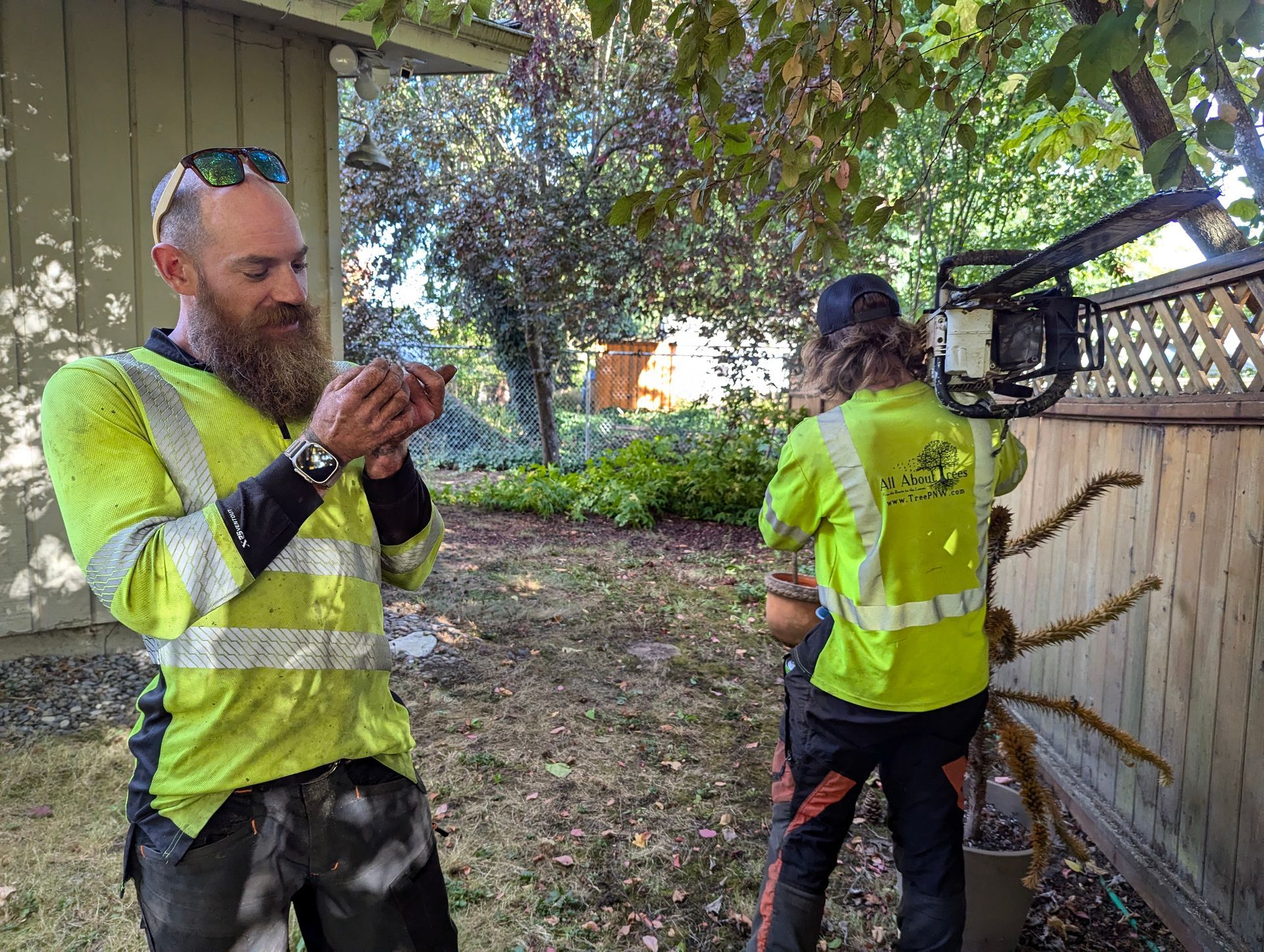 Two workers in neon safety vests standing outdoors beside a fence and lifting equipment under trees
