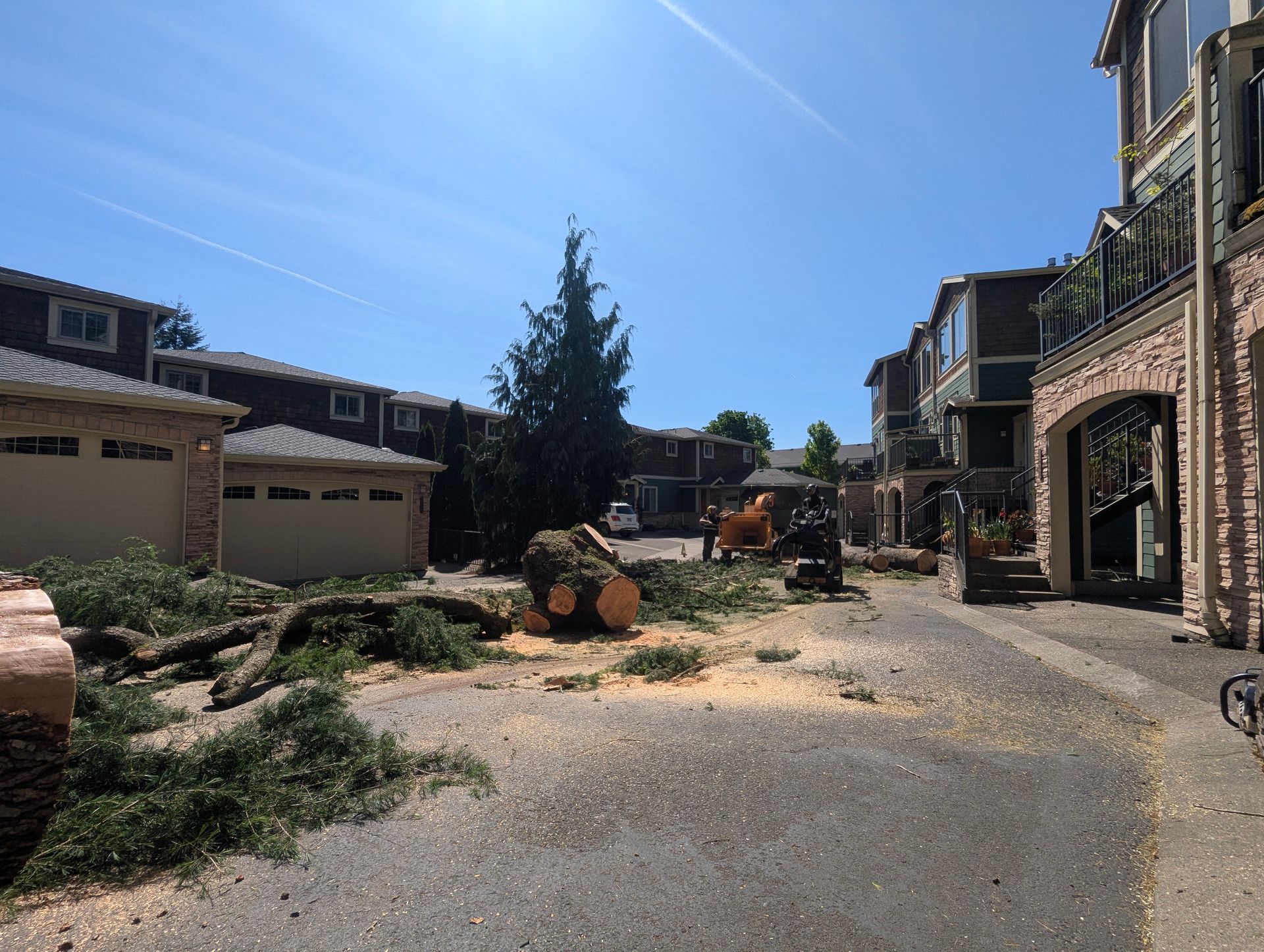 Residential driveway with construction debris, excavator, and a pile of cut tree logs beside houses under a blue sky