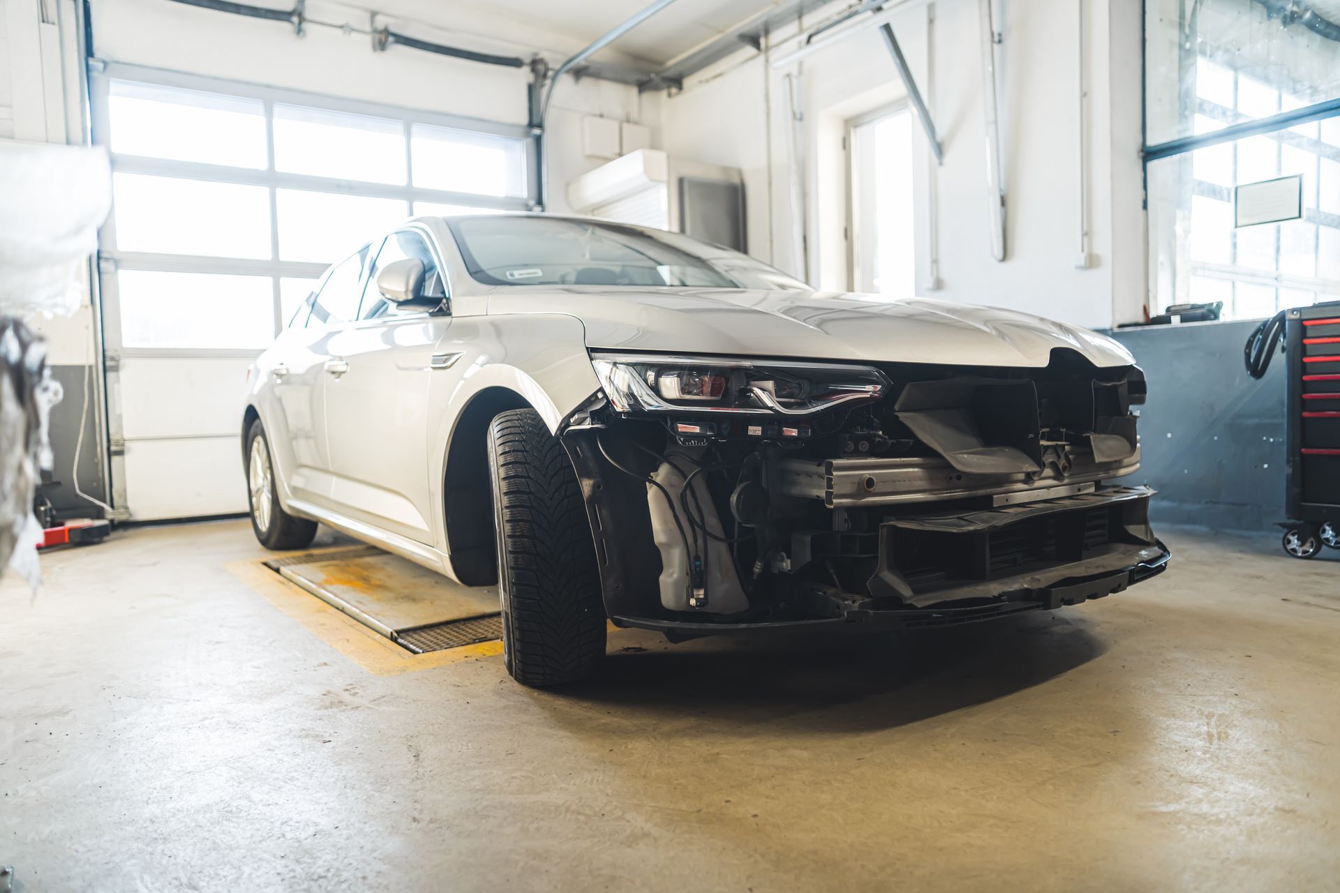 Damaged white car in a bright auto repair garage, front bumper removed and front end exposed