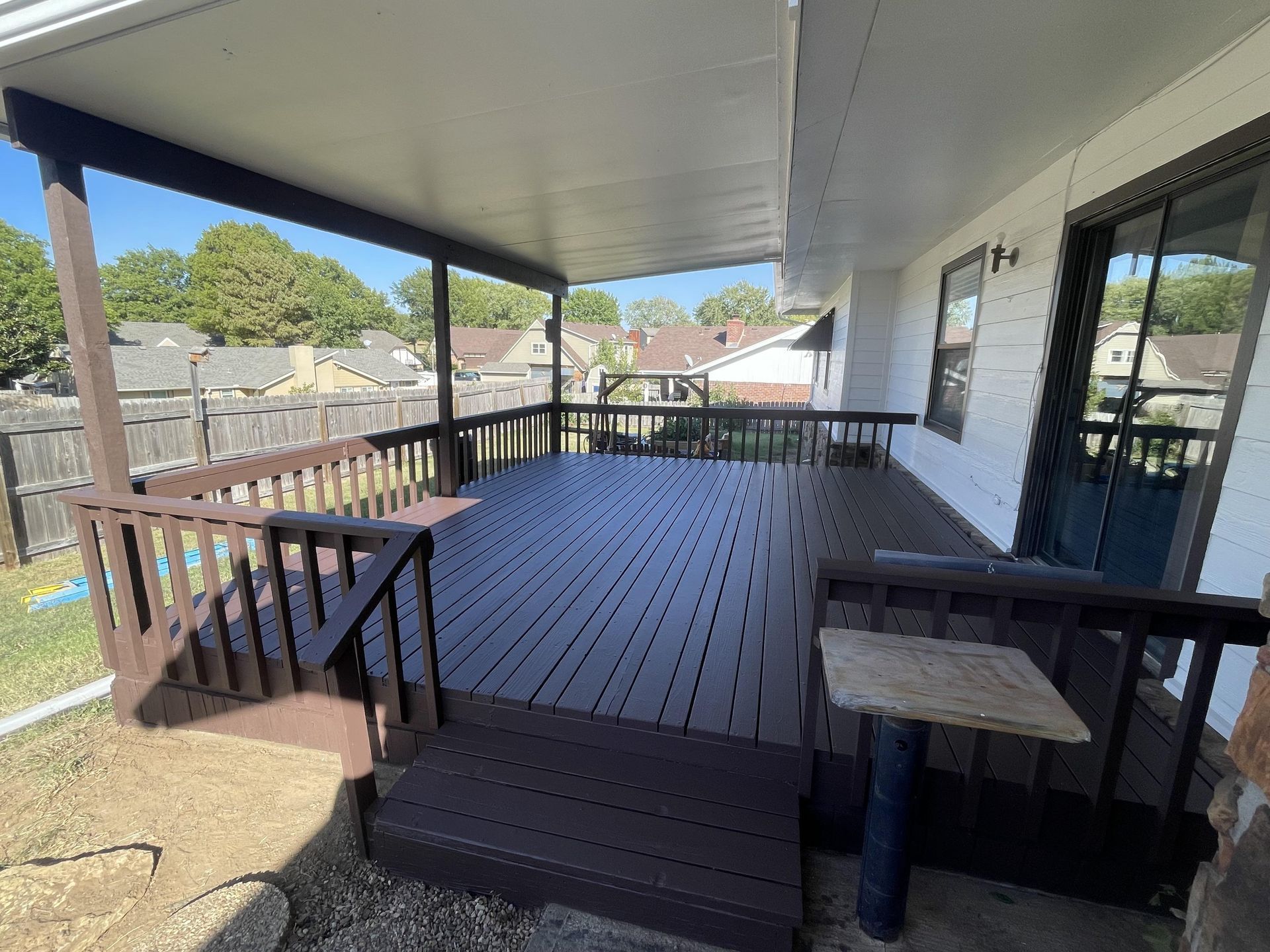 A raised, dark-stained wooden deck with railings, sheltered by a covered patio roof, next to a white house.