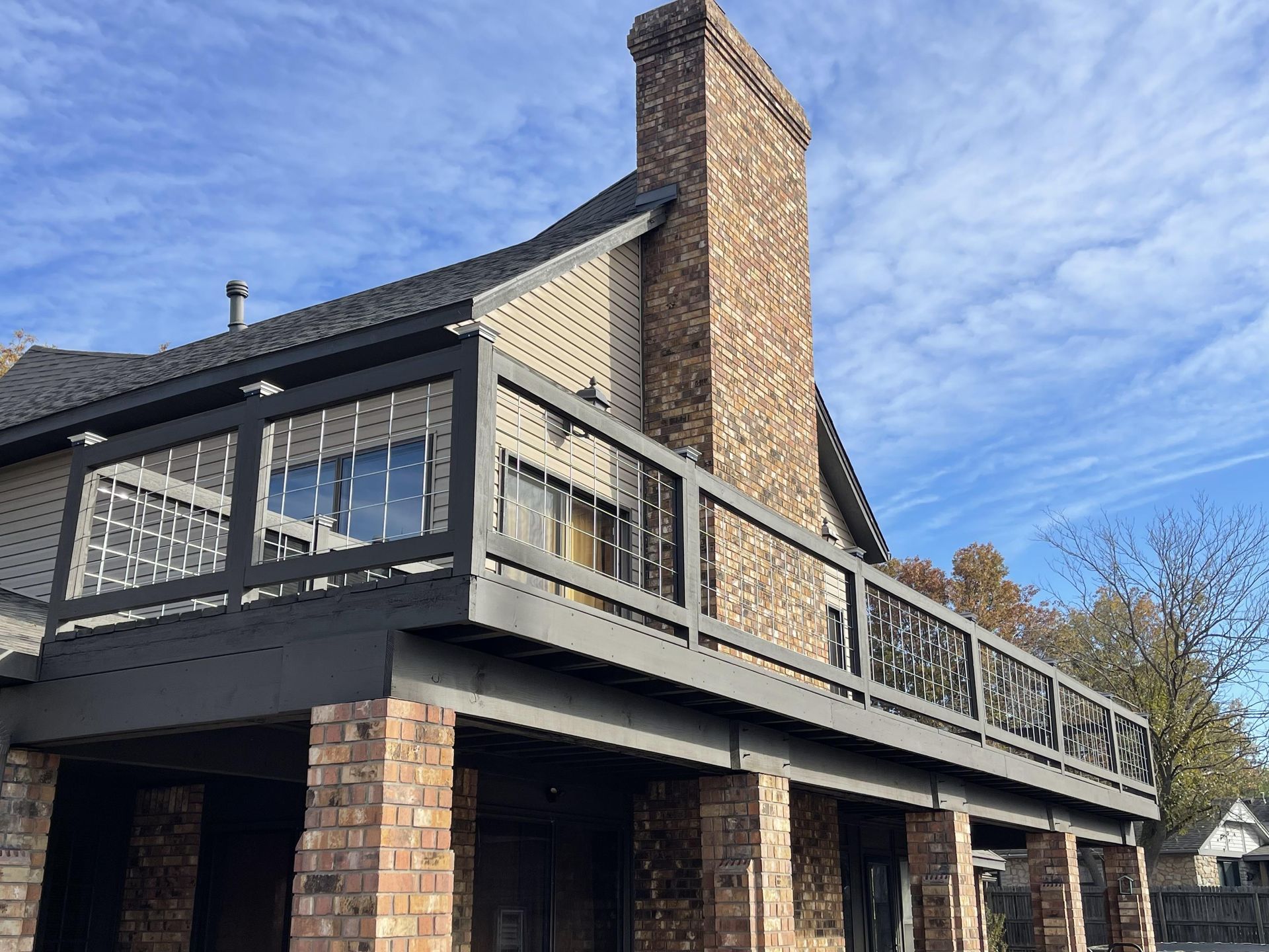 A tan brick house with a tall chimney and a large, dark-painted wooden deck under a bright blue, cloudy sky.