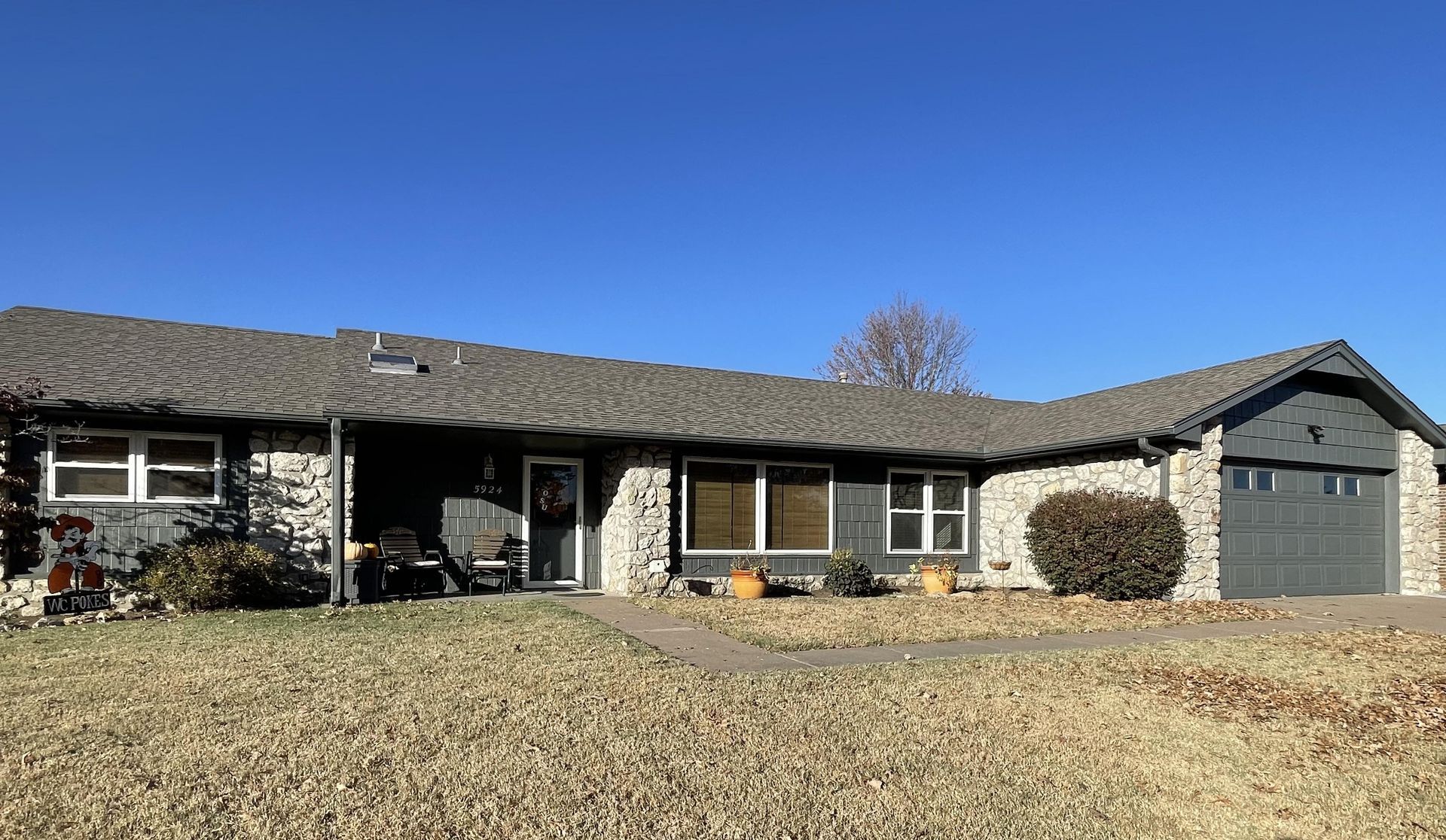 A single-story suburban house with a gray roof, stone-accented facade, and a attached garage under a bright blue sky.