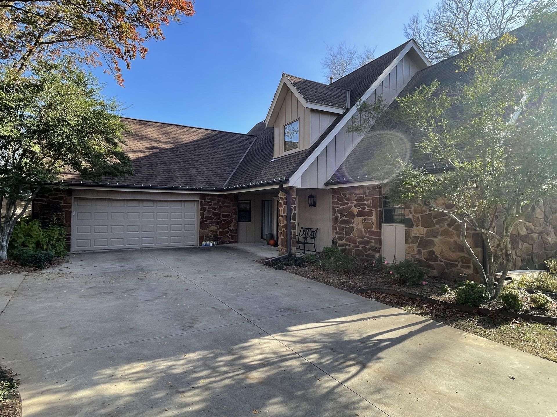 A two-story suburban home with a stone and wood exterior, a two-car garage, a concrete driveway, and surrounding trees.