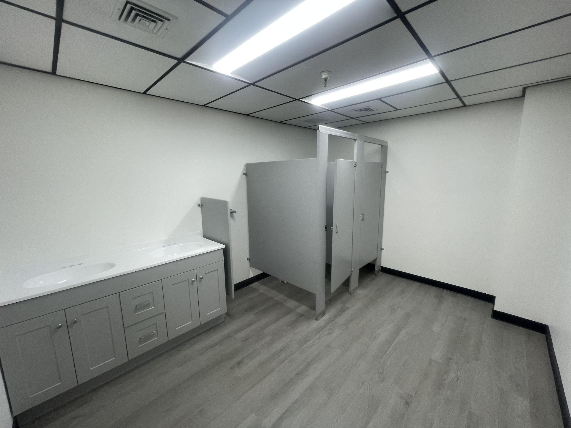 A minimalist public restroom with light gray vanity cabinets, wood-look flooring, white walls, and two stall partitions.