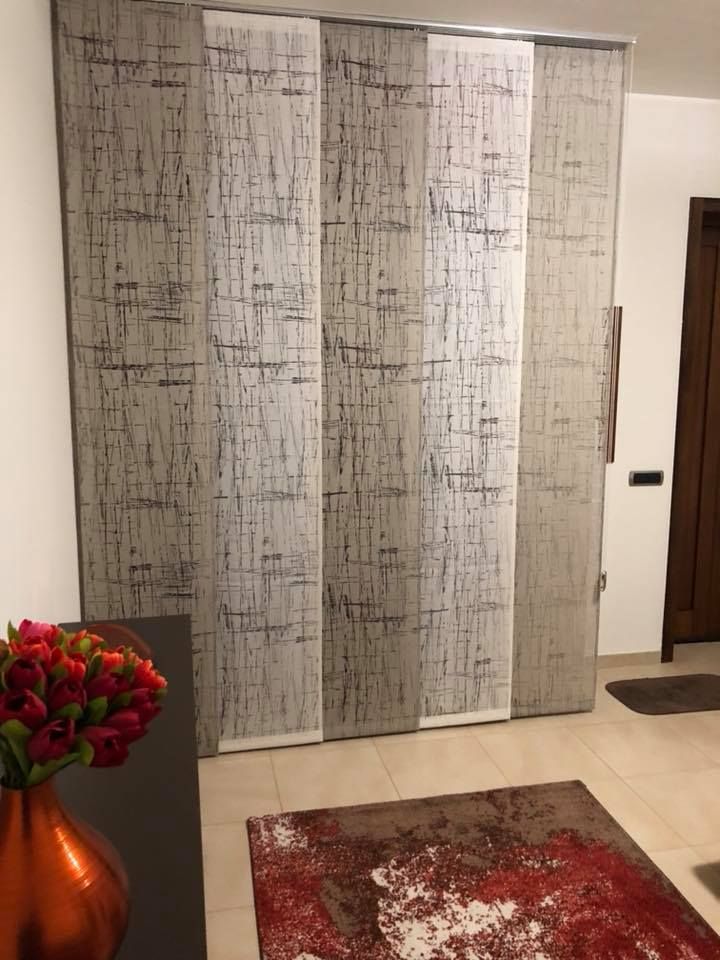 Soggiorno con alte tende grigie a fantasia, tappeto rosso e vaso di rame con fiori rossi.