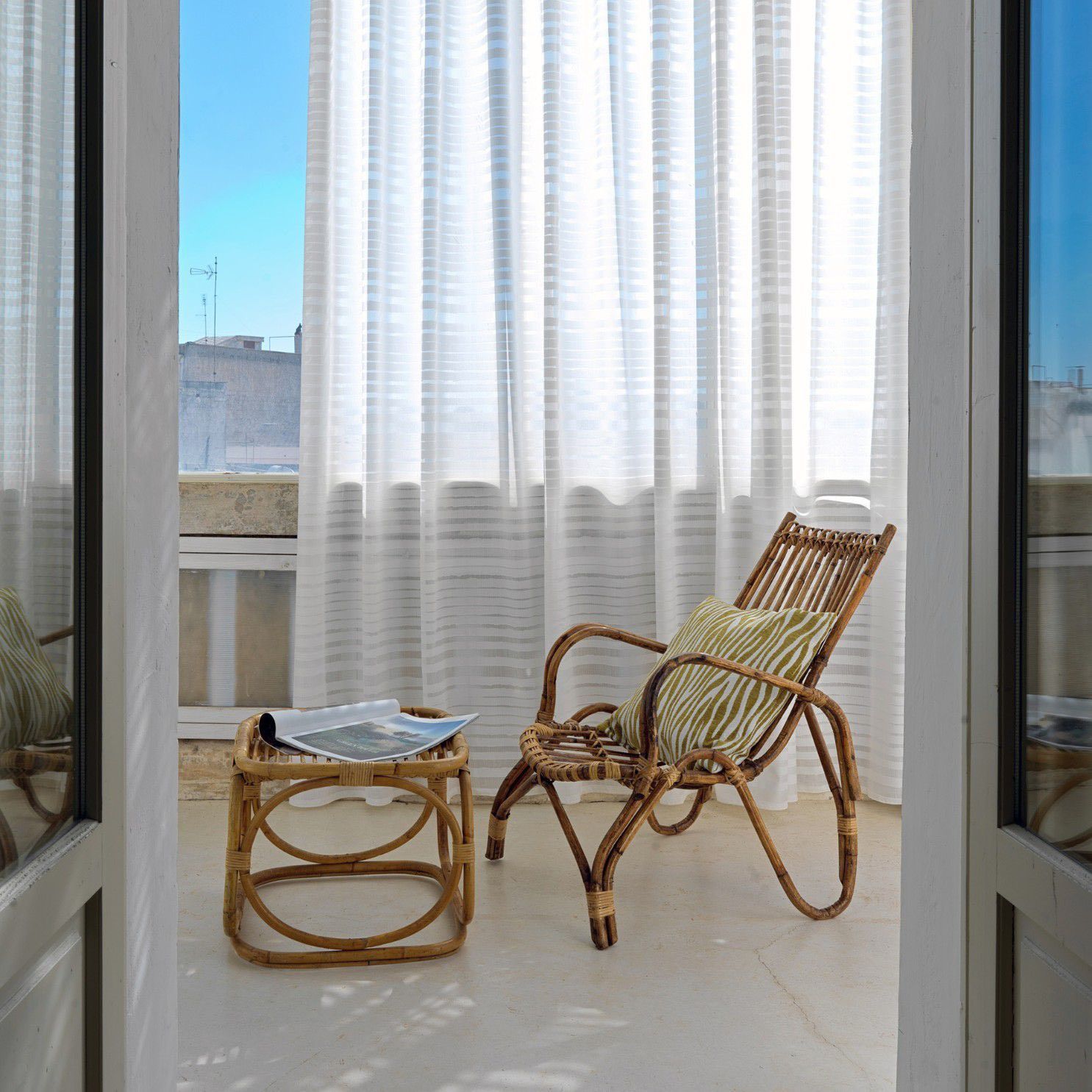 Balcone soleggiato con sedia in vimini, tavolino abbinato e tende bianche trasparenti accanto alle porte aperte.