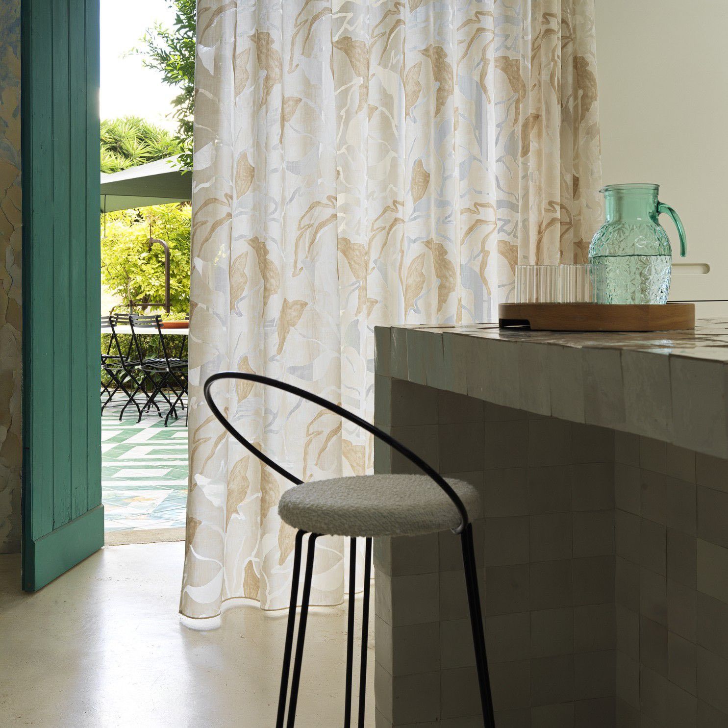 Si apre una porta su un patio soleggiato, con una tenda color crema, una sedia nera e un bancone grigio con una brocca di vetro.