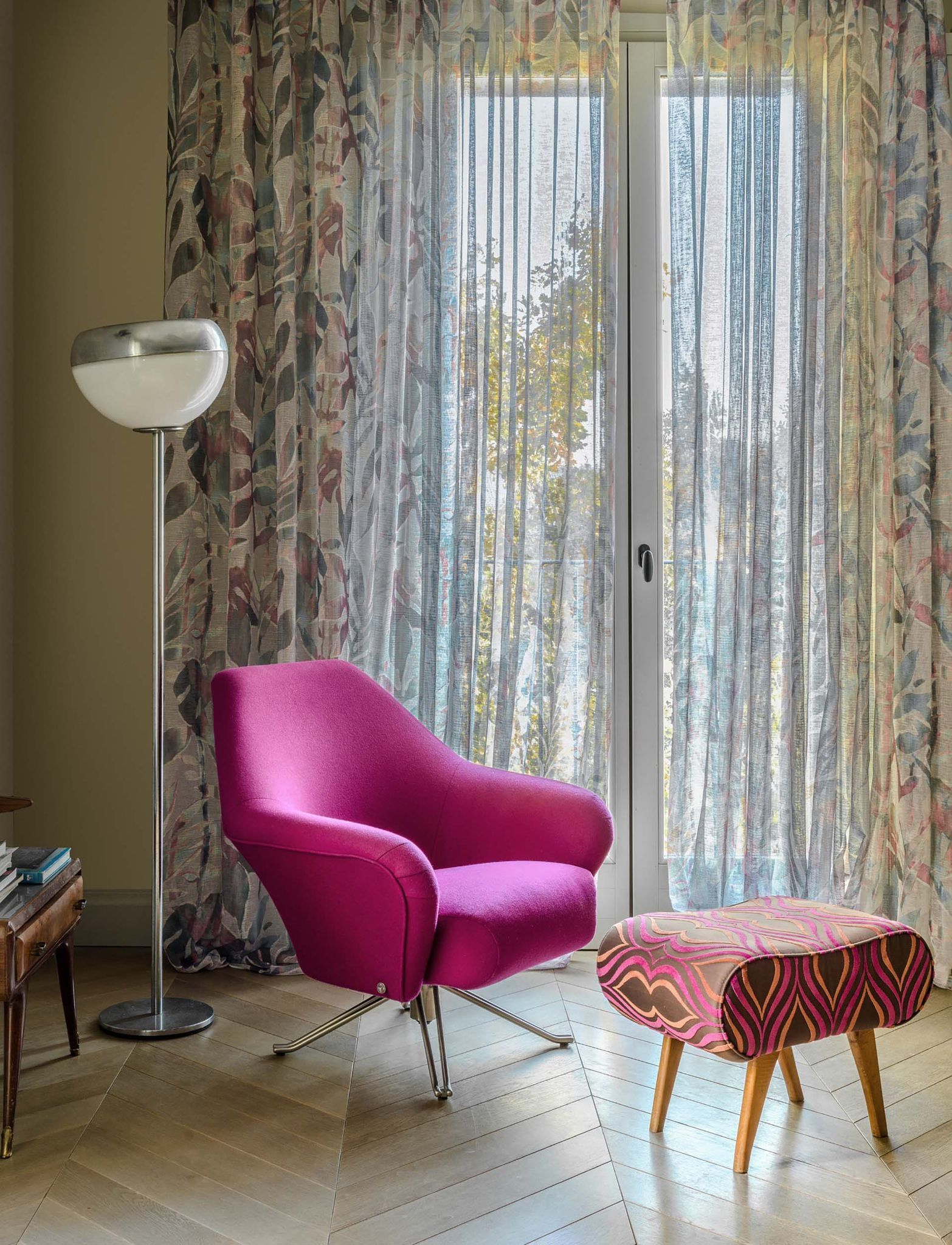 In un accogliente soggiorno, una poltrona rosa e un pouf a fantasia sono posizionati accanto a una finestra con tende trasparenti.