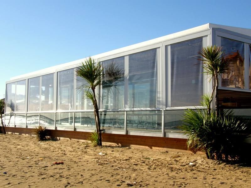 Moderno padiglione in vetro fronte mare con struttura bianca, palme e primo piano sabbioso sotto un cielo azzurro limpido.