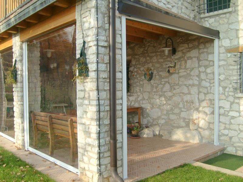 Veranda in pietra con finestre a zanzariera, pianta sospesa e panchina in legno su un piccolo patio.