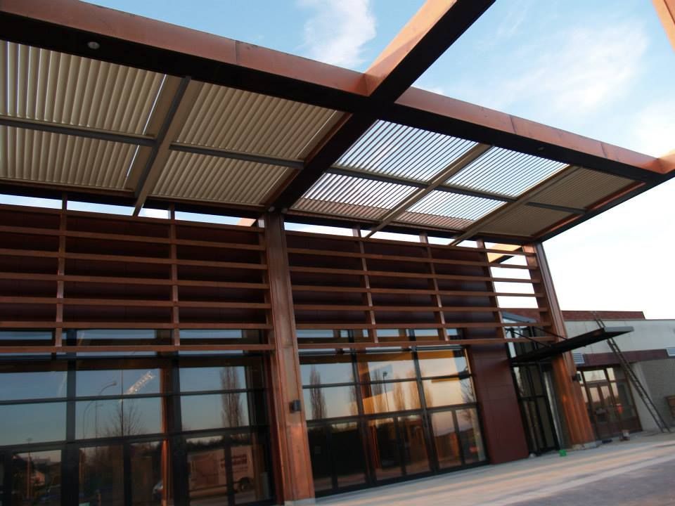 Ingresso di un edificio moderno con listelli di legno, pareti in vetro e una tettoia in lamiera ondulata sotto un cielo azzurro.