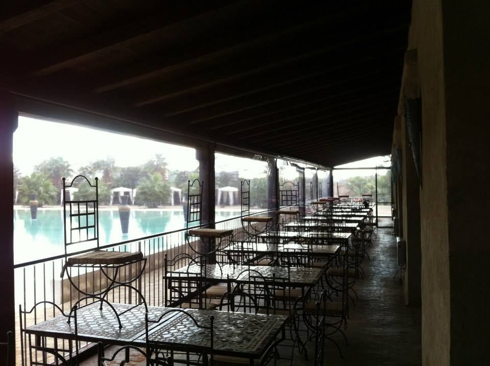 Patio esterno con tavoli e sedie in metallo a bordo piscina, sotto una tettoia.
