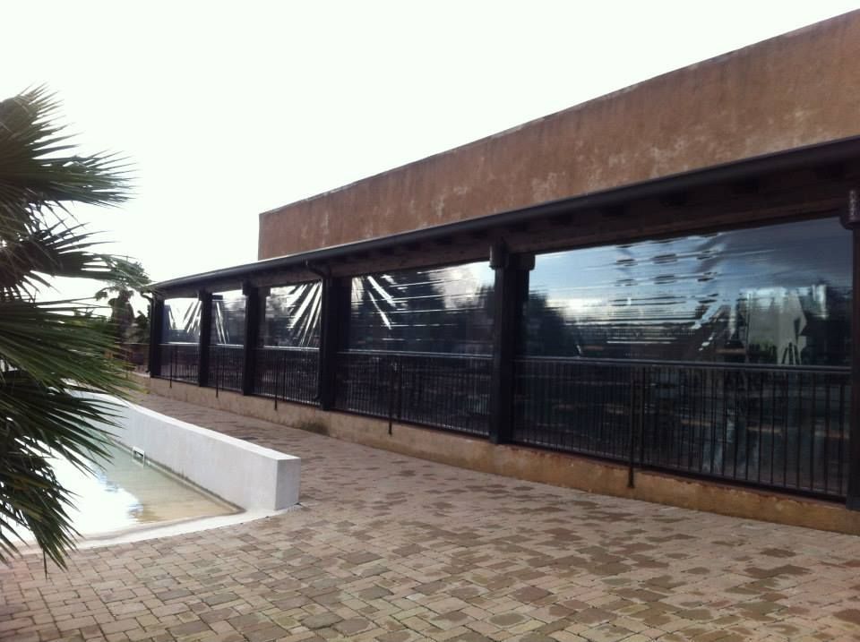 Edificio moderno con facciata in vetro e tetto marrone, visto da un cortile pavimentato con foglie di palma sulla sinistra.