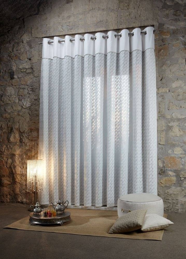Stanza con pareti in pietra, tende bianche trasparenti, luce calda di una lampada e un cuscino sul pavimento.