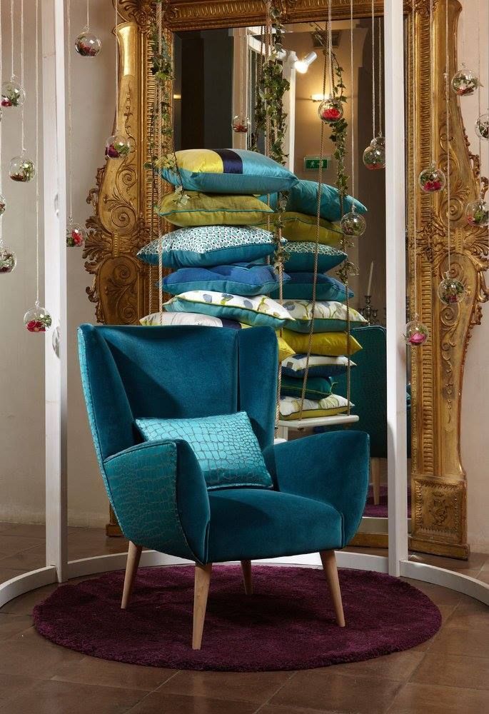 Poltrona color verde acqua con cuscini decorativi impilati davanti a uno specchio ornato con cornice dorata.