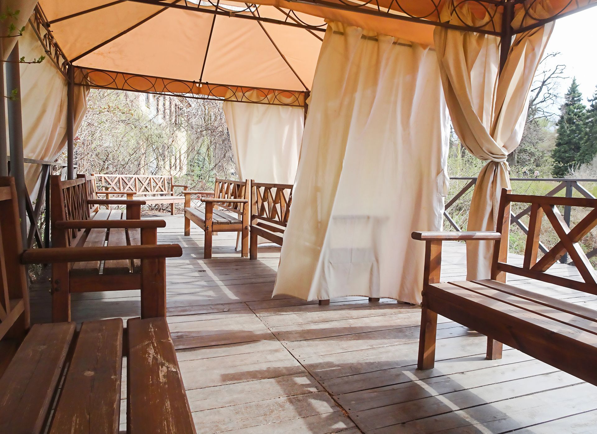 Gazebo in legno con tende, panchine e tavoli affacciato su un paesaggio boschivo.