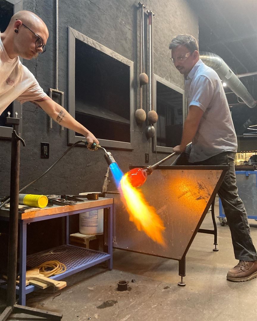 Two people in a glassblowing studio, one holding a rod with molten glass, other using a torch. Flames visible.