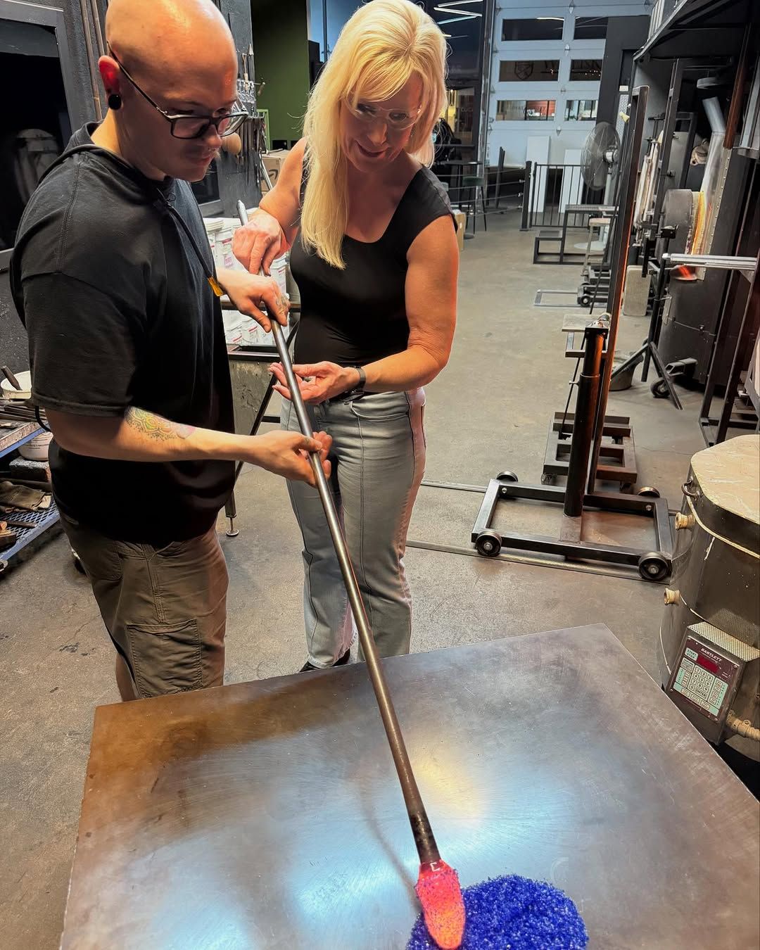 Man and woman working with molten glass in a workshop, using a long rod and shaping tools.