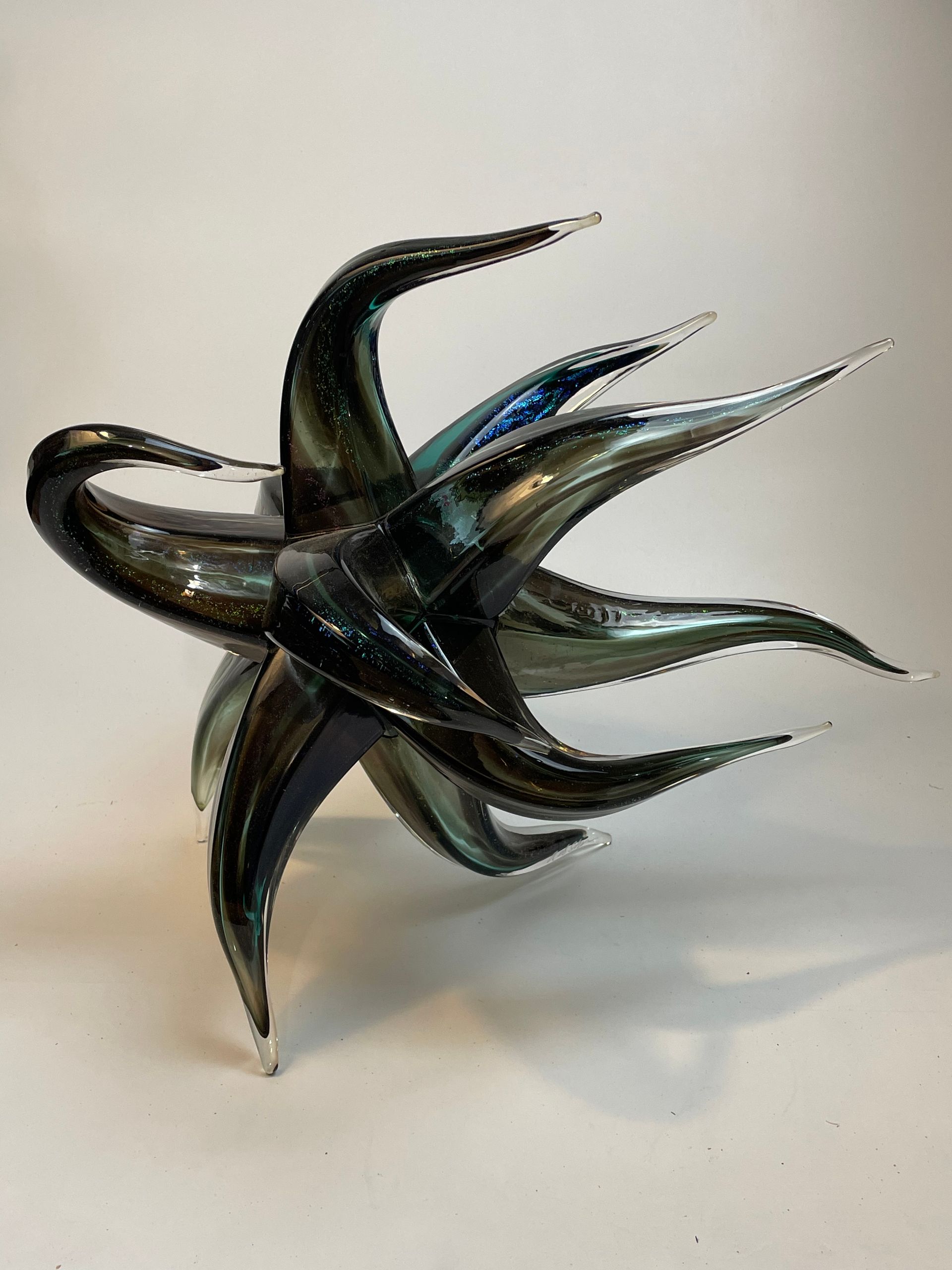 Sculpted glass art with swirling, spiky dark blue and green shapes against a white background.