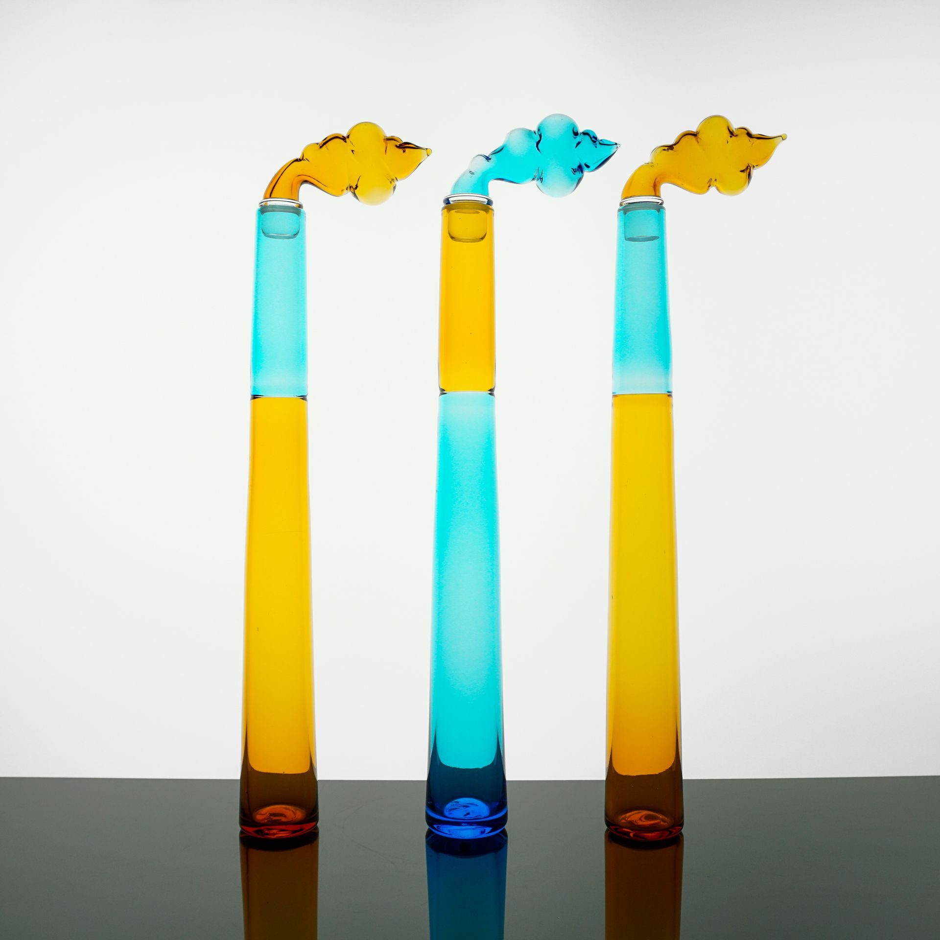 Three glass sculptures resembling smokestacks, blue and yellow with cloud-shaped tops, on a reflective surface.