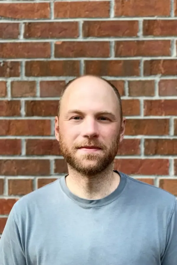 Man with a beard and shaved head in a gray shirt, standing in front of a brick wall.