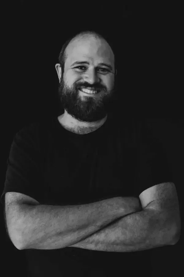 Man with beard smiling, arms crossed, wearing a dark t-shirt, against a black background.