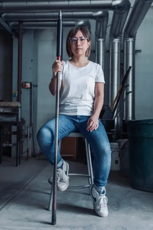 Woman seated, holding a long tool. Wearing white shirt, jeans, and sneakers. Industrial setting with pipes.