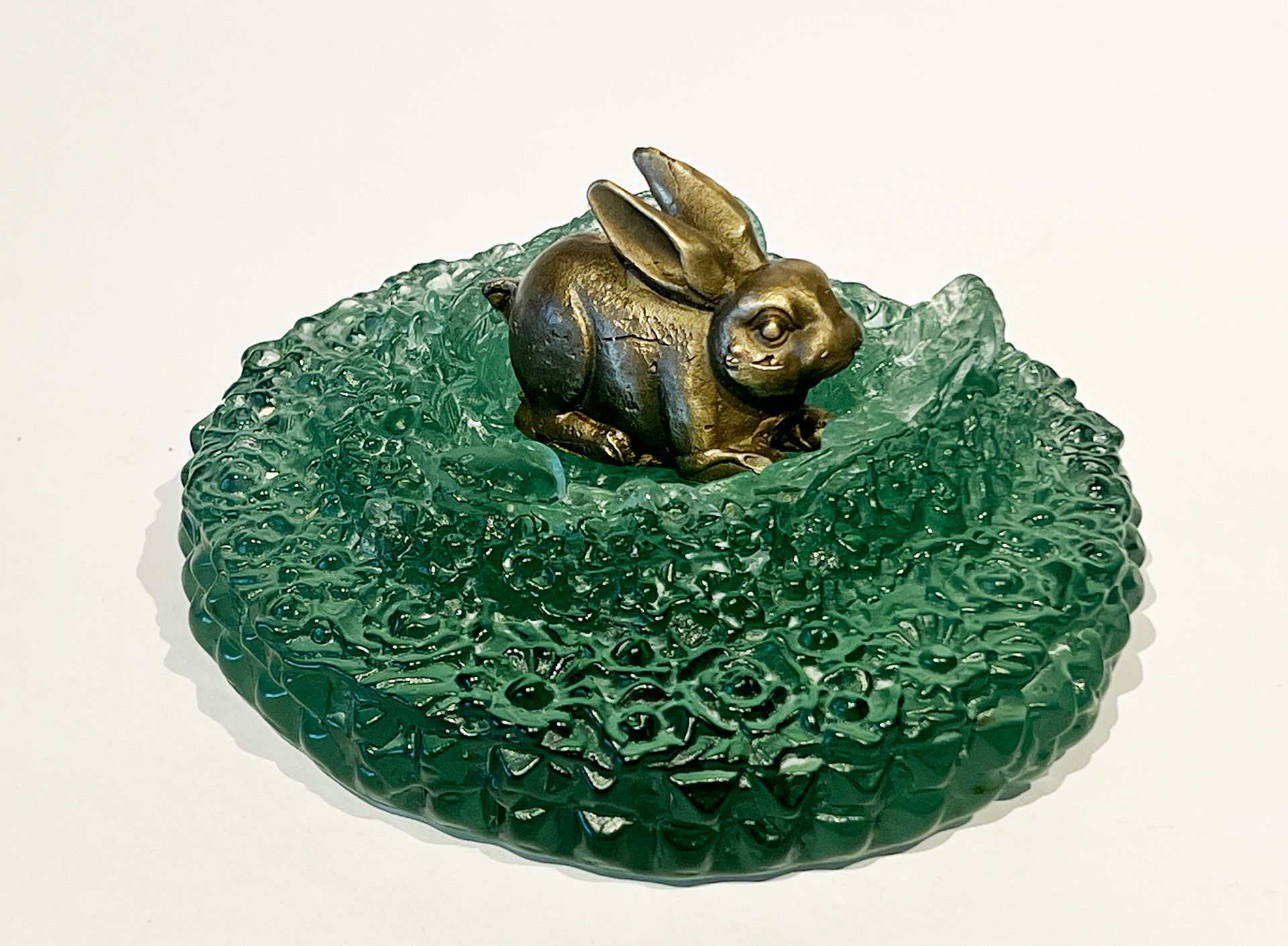 Bronze rabbit sits in a bed of textured, emerald green glass on a white surface.
