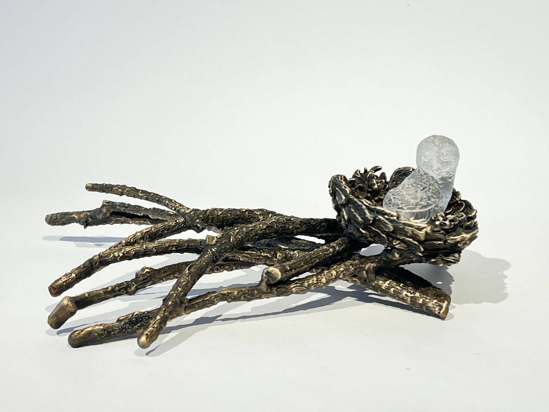 Bronze sculpture of a bird nest with branches. A frosted glass bird sits in the center, all against a white background.