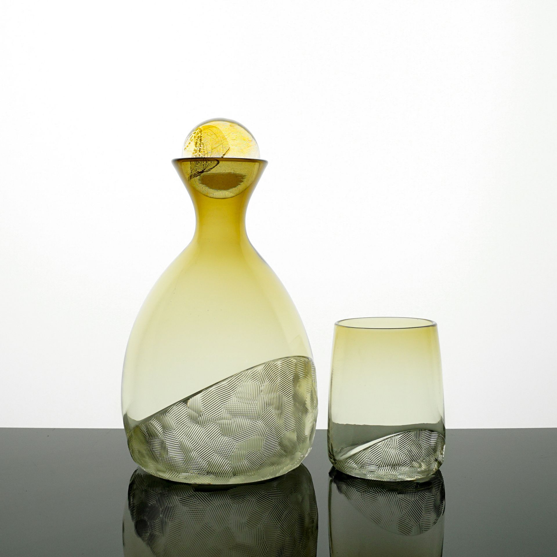 Yellow glass decanter and tumbler, with textured base, on a reflective surface.