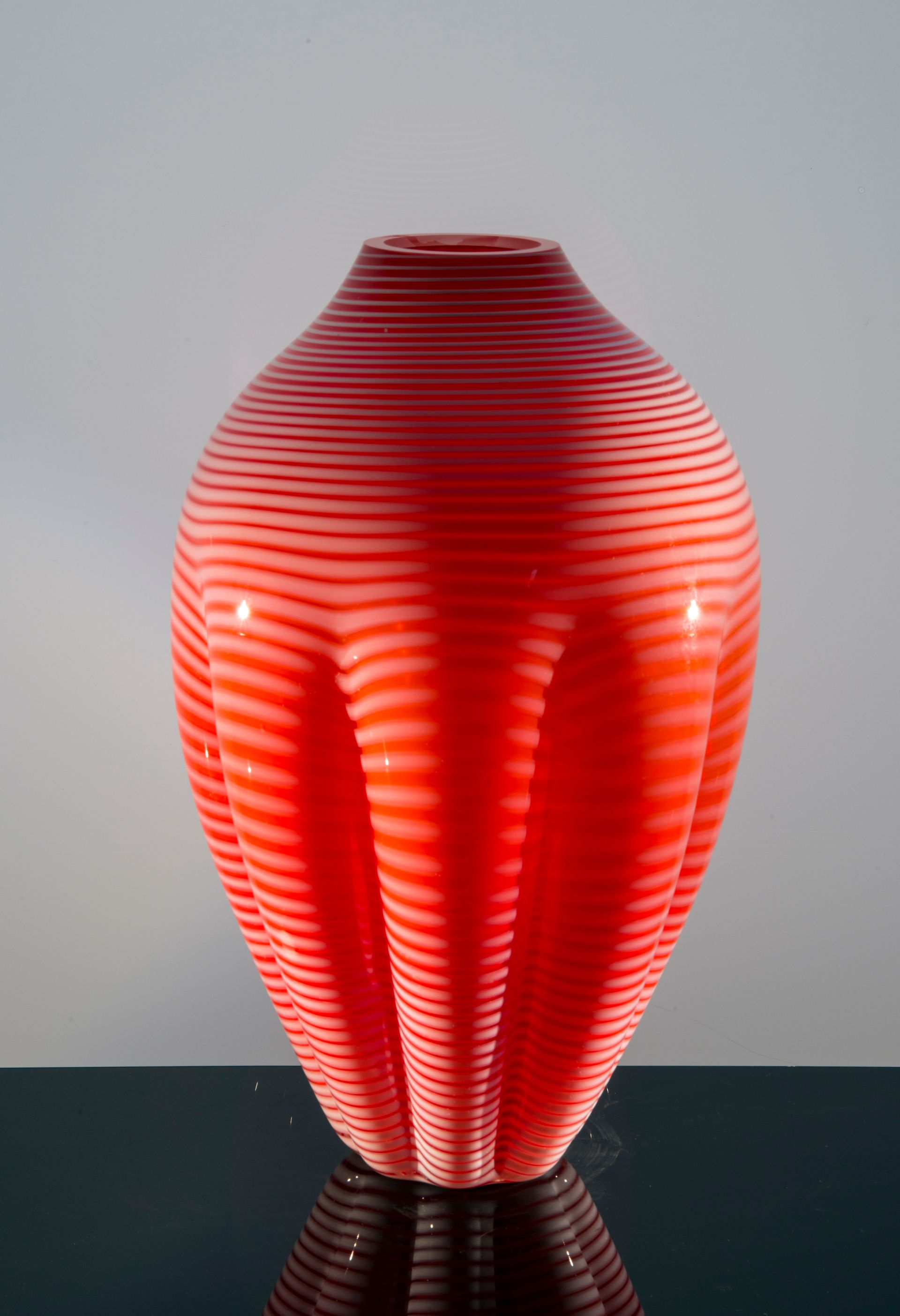 Red glass vase with horizontal stripes, on reflective surface.