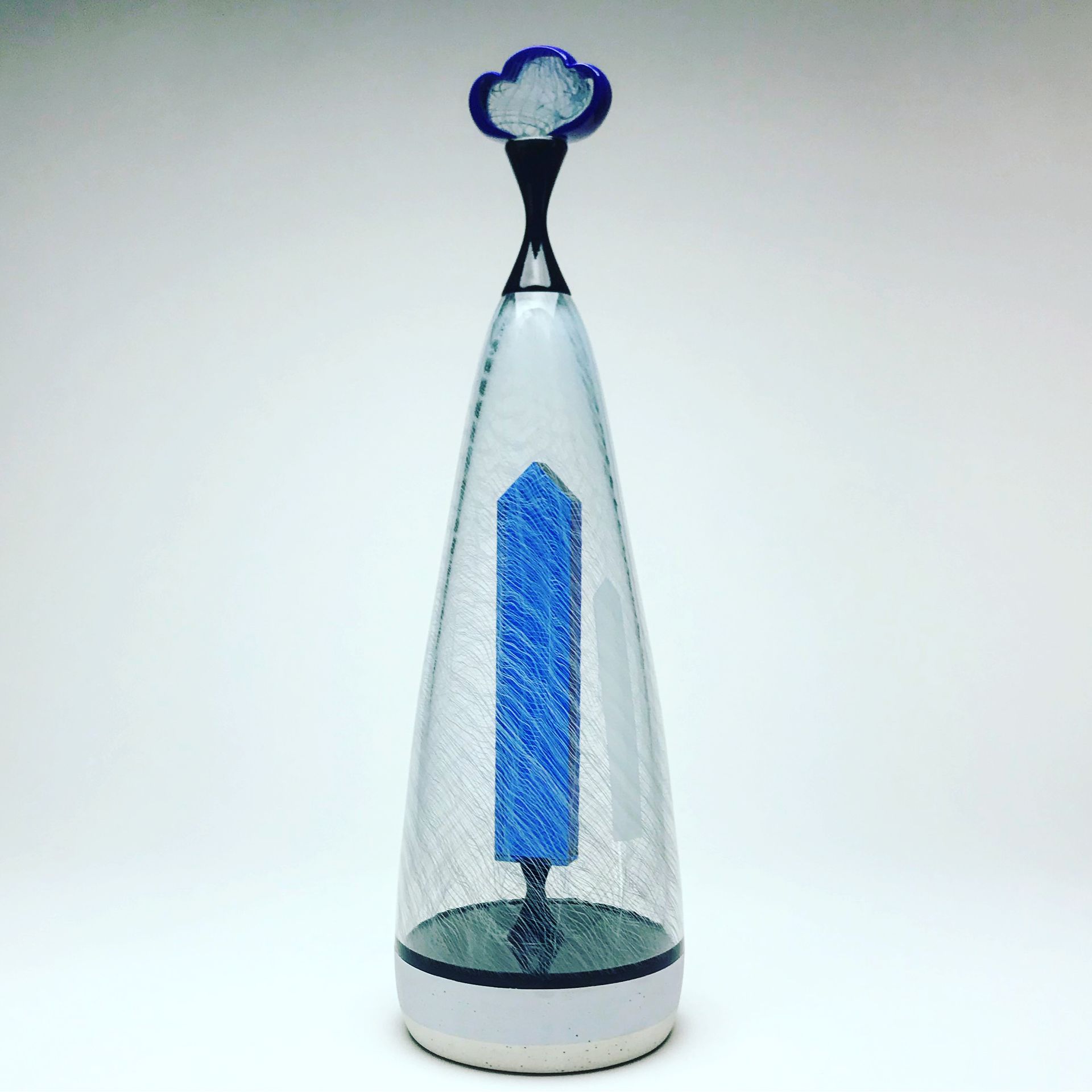 A clear glass enclosed sculpture with a blue pillar and a cloud design on top; against a white backdrop.